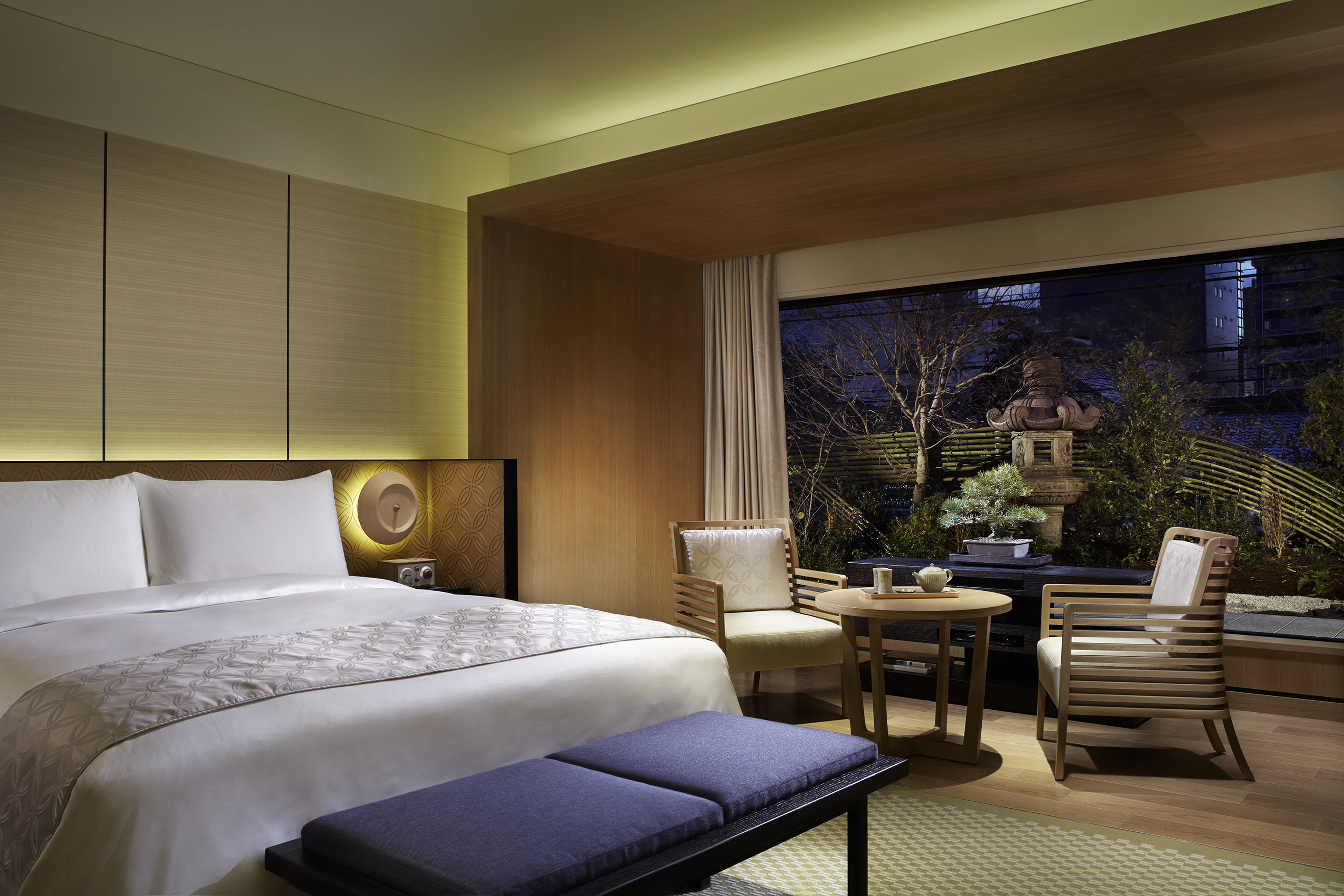 A cosy suite lit up at night with a comfy bed and pale wood furniture, with a view of gardens through the large window