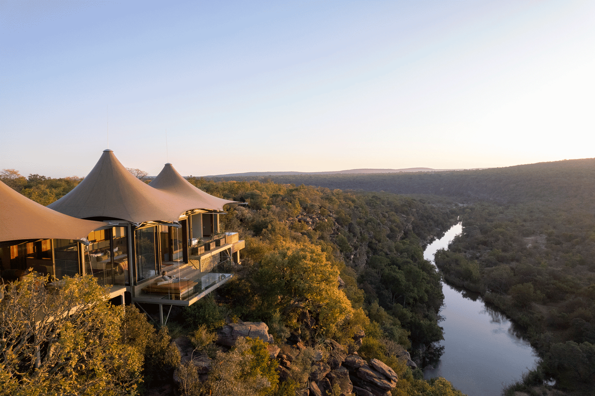 Luxury safari-style lodge perched on a cliff overlooking a winding river and expansive landscape at sunset.