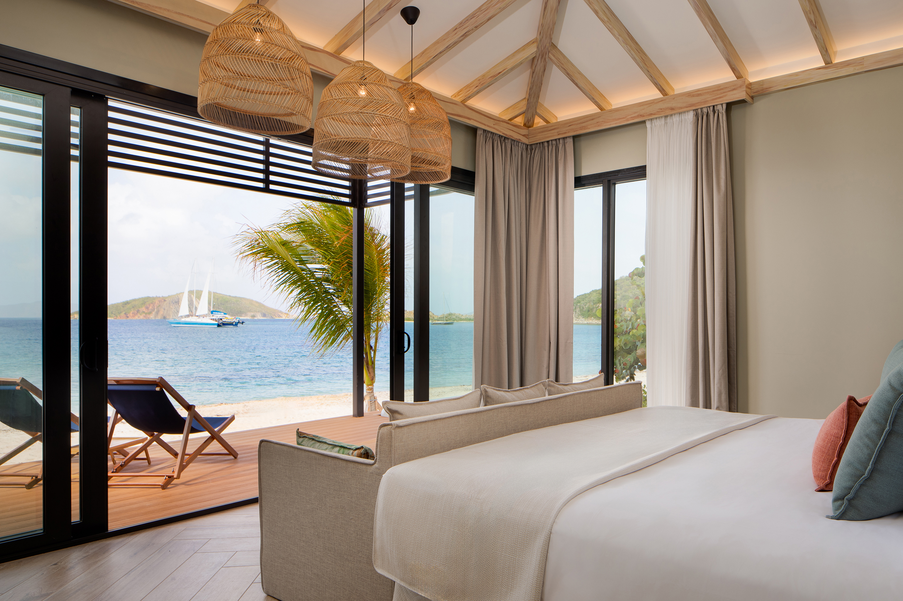 Airy bedroom with white bedding and rattan light shades facing floor to ceiling glass doors overlooking the ocean with a boat drifting by