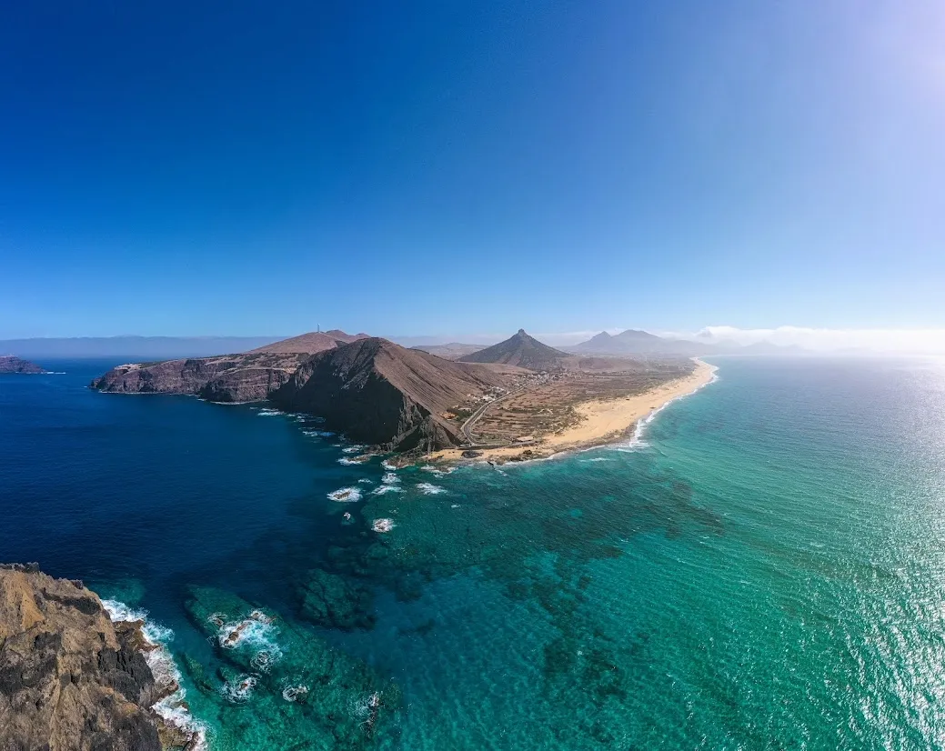 Aerial view of a rugged coastal peninsula with turquoise and deep blue waters, a sandy shoreline, and mountain peaks under a clear blue sky.
