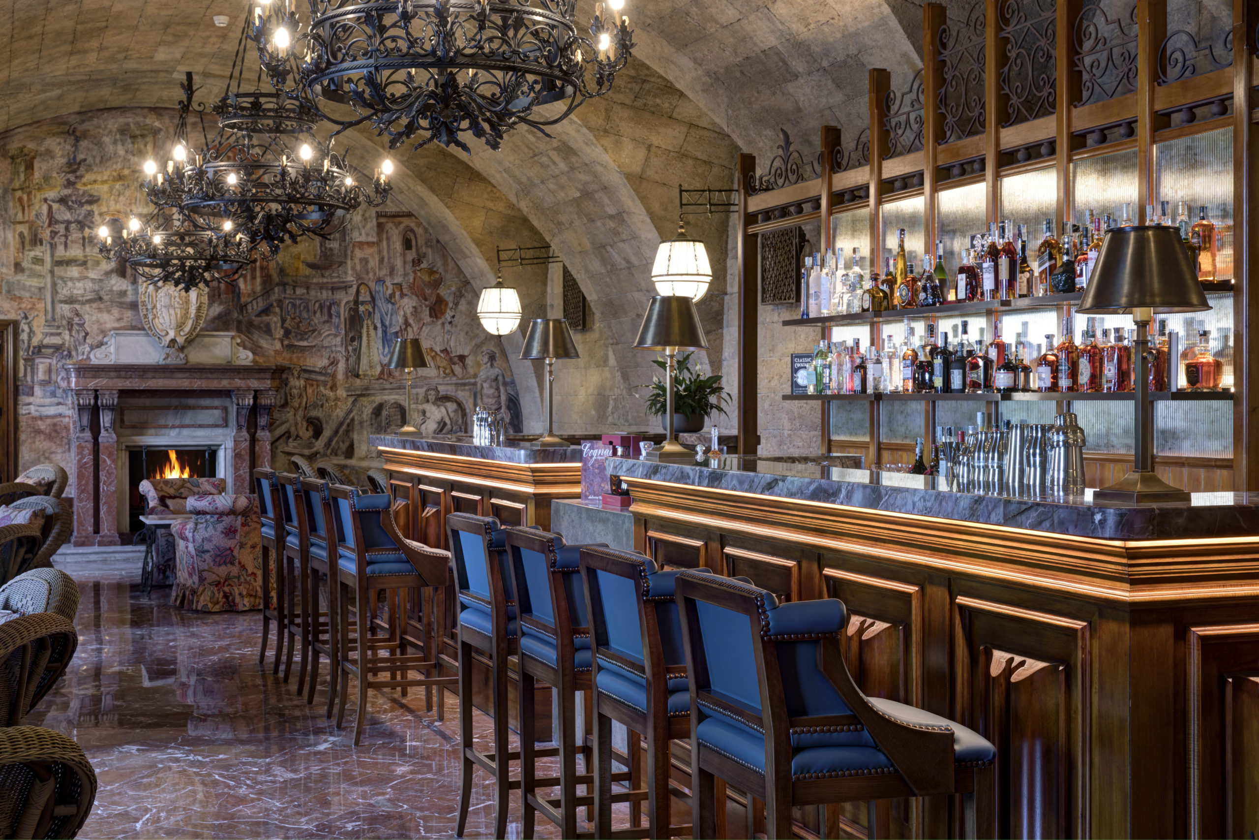 An opulent bar with a wooden counter, blue upholstered stools, and a grand stone fireplace, under an arched ceiling with chandeliers and mural walls.