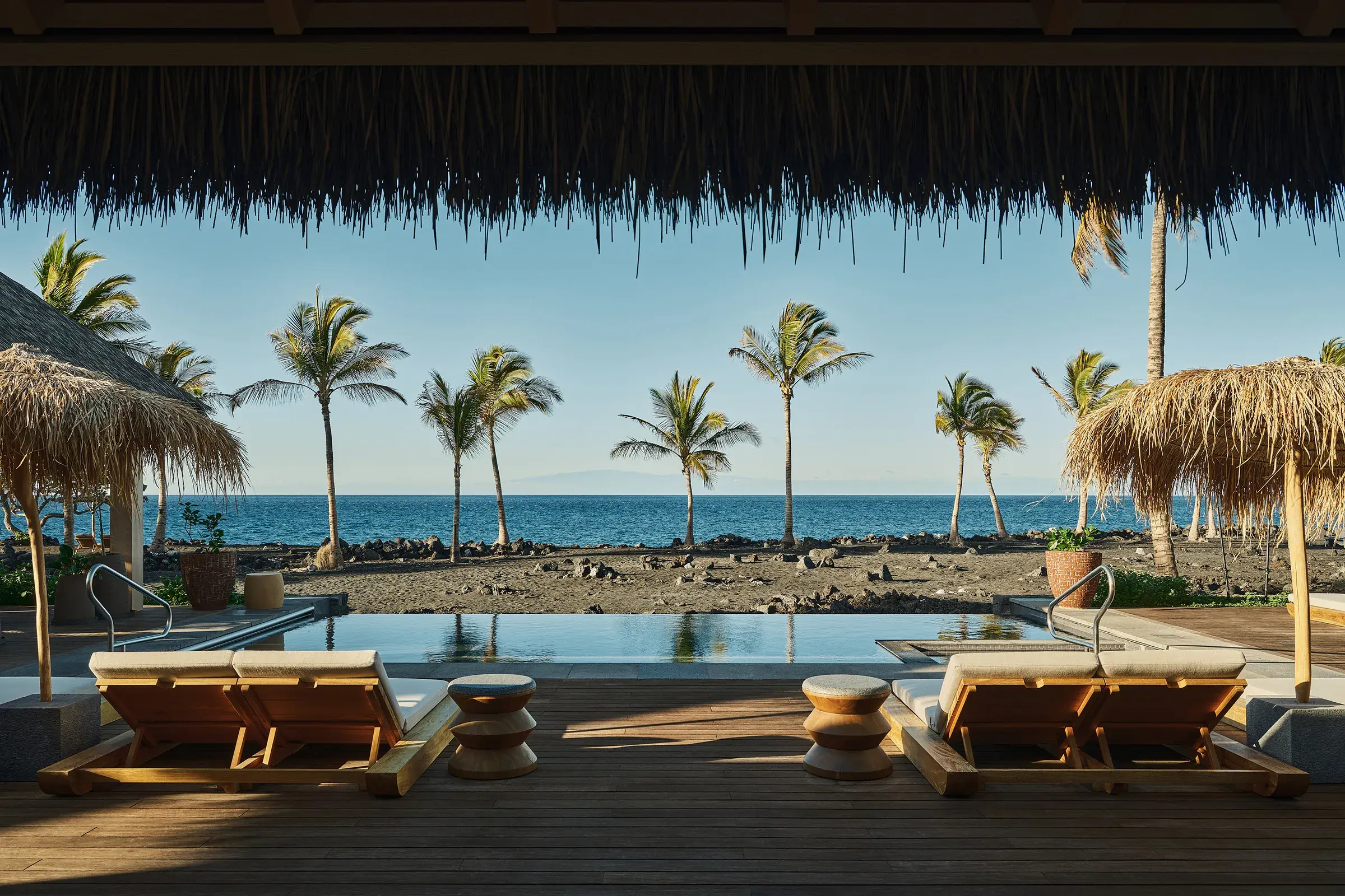 The pool of a Signature Suite Kumukea at Rosewood Kona Village with sun loungers facing palm trees and the ocean, framed by a straw roof
