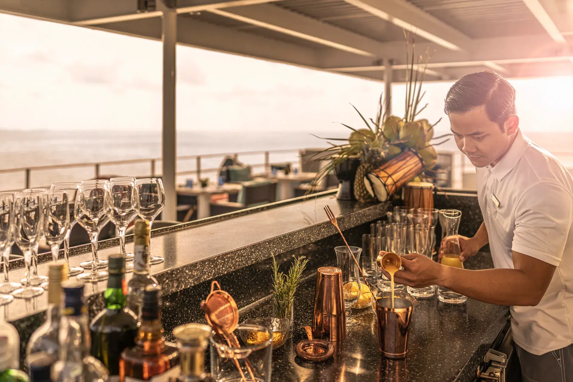 A bartender making a drink aboard Four Seasons Explorer Palau surrounded by glasses and bottles with a view of the ocean behind