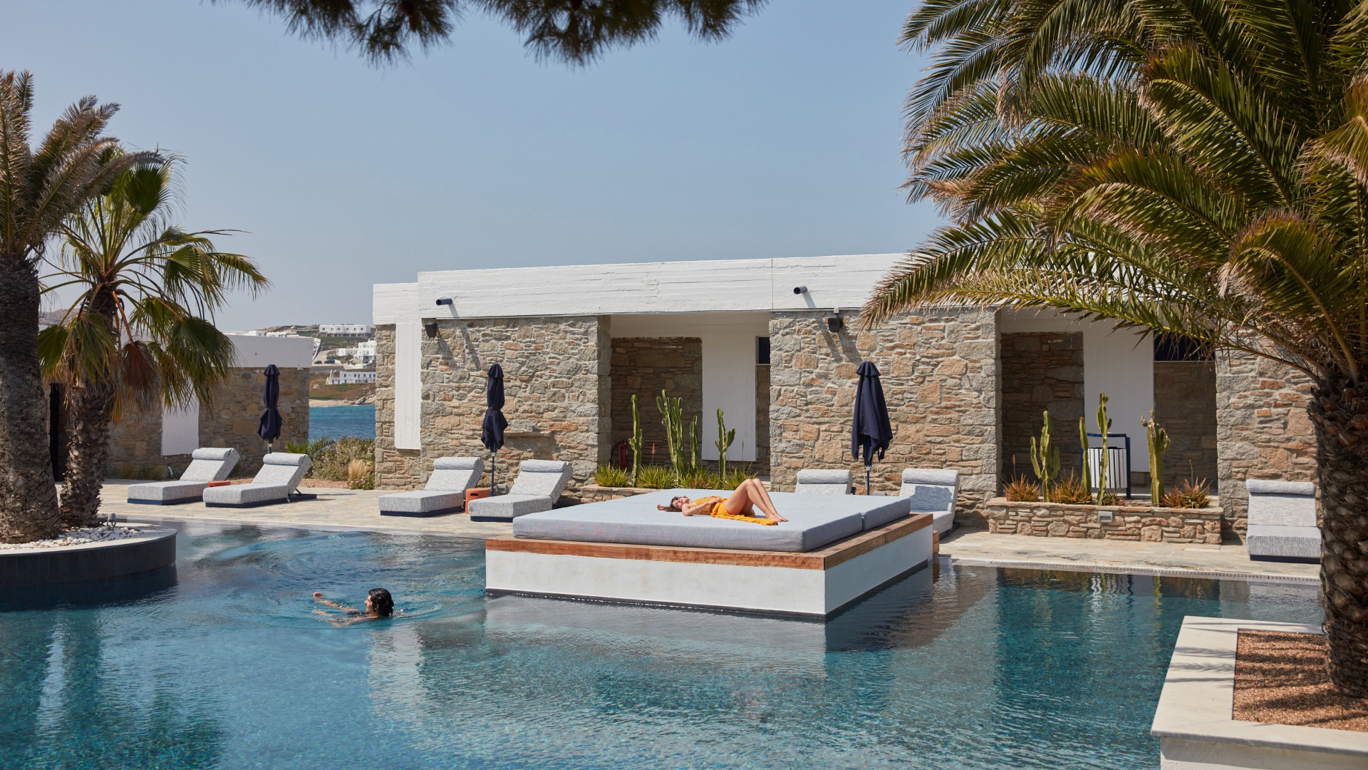 The large outdoor pool are of Mykonos Theoxenia surrouned by loungers and palms with one person swimming and one lounging on a large square lounger
