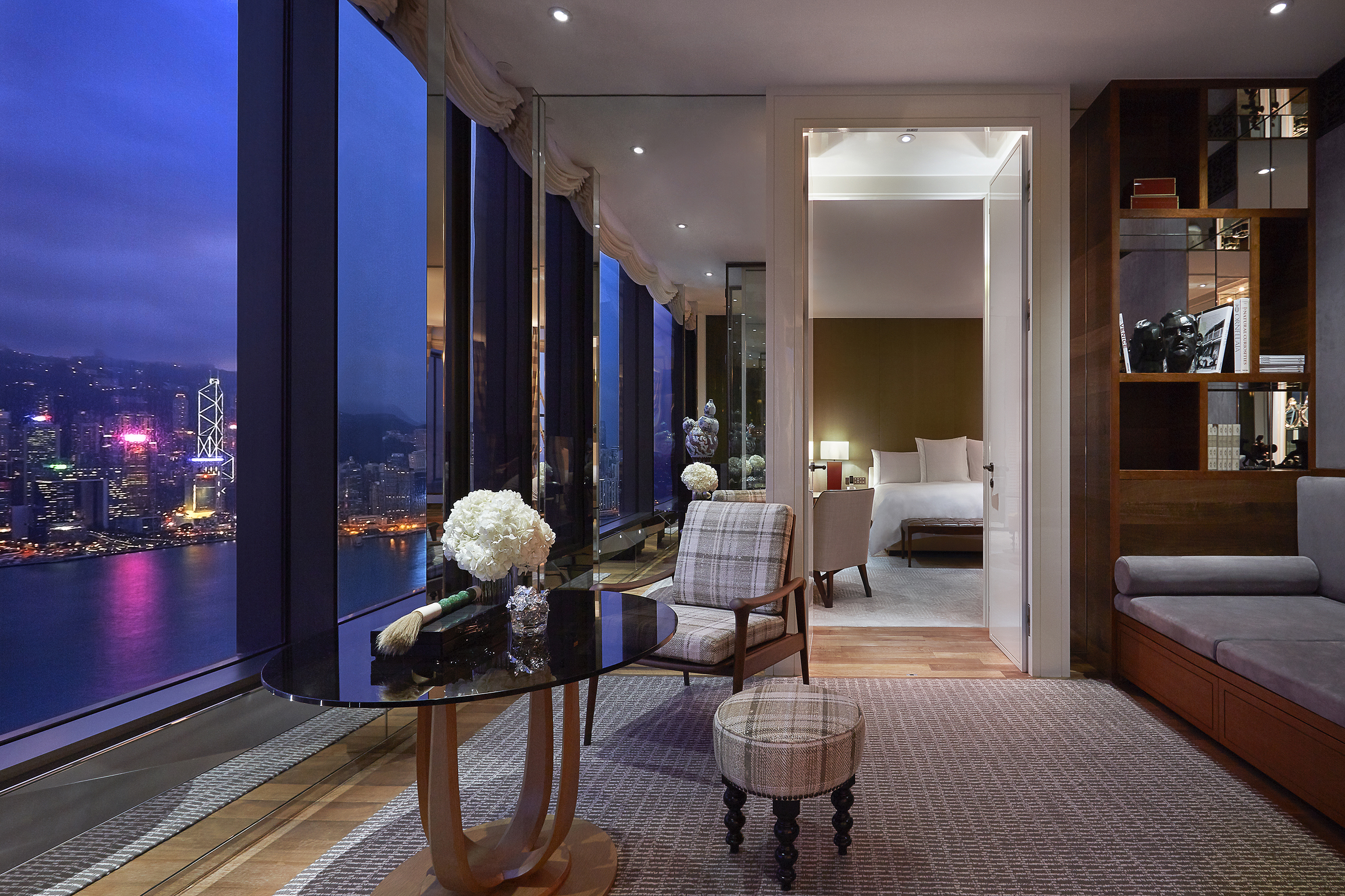 A Manor Suite at Rosewood Hong Kong with a single table and chair set before the floor-to-ceiling windows with city views, decorated shelving and a glimpse of the bedroom beyond