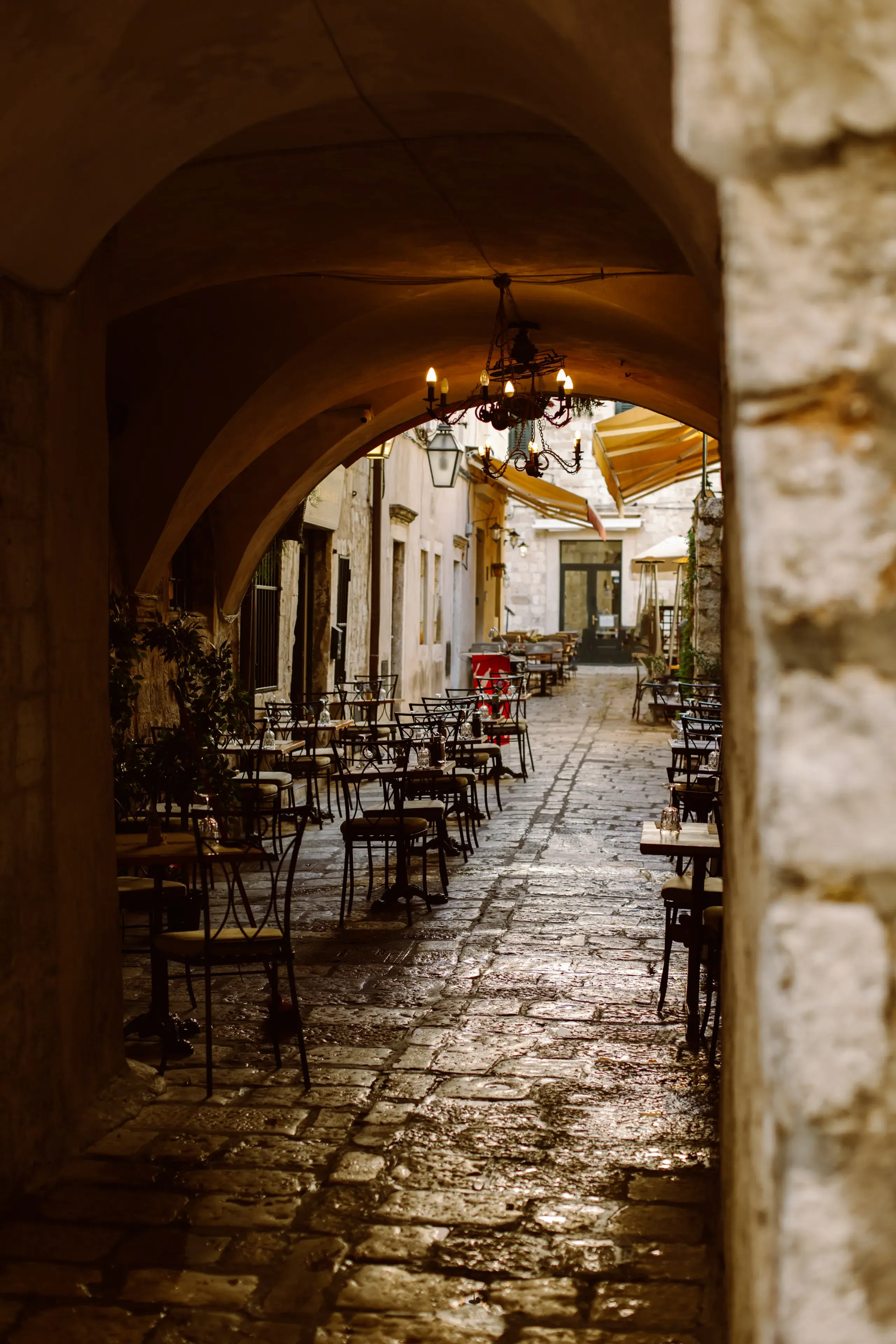 A dimly lit alley in Dubrovnik leading to a restaurant with wooden tables and chairs beneath a stone arch with a chandelier