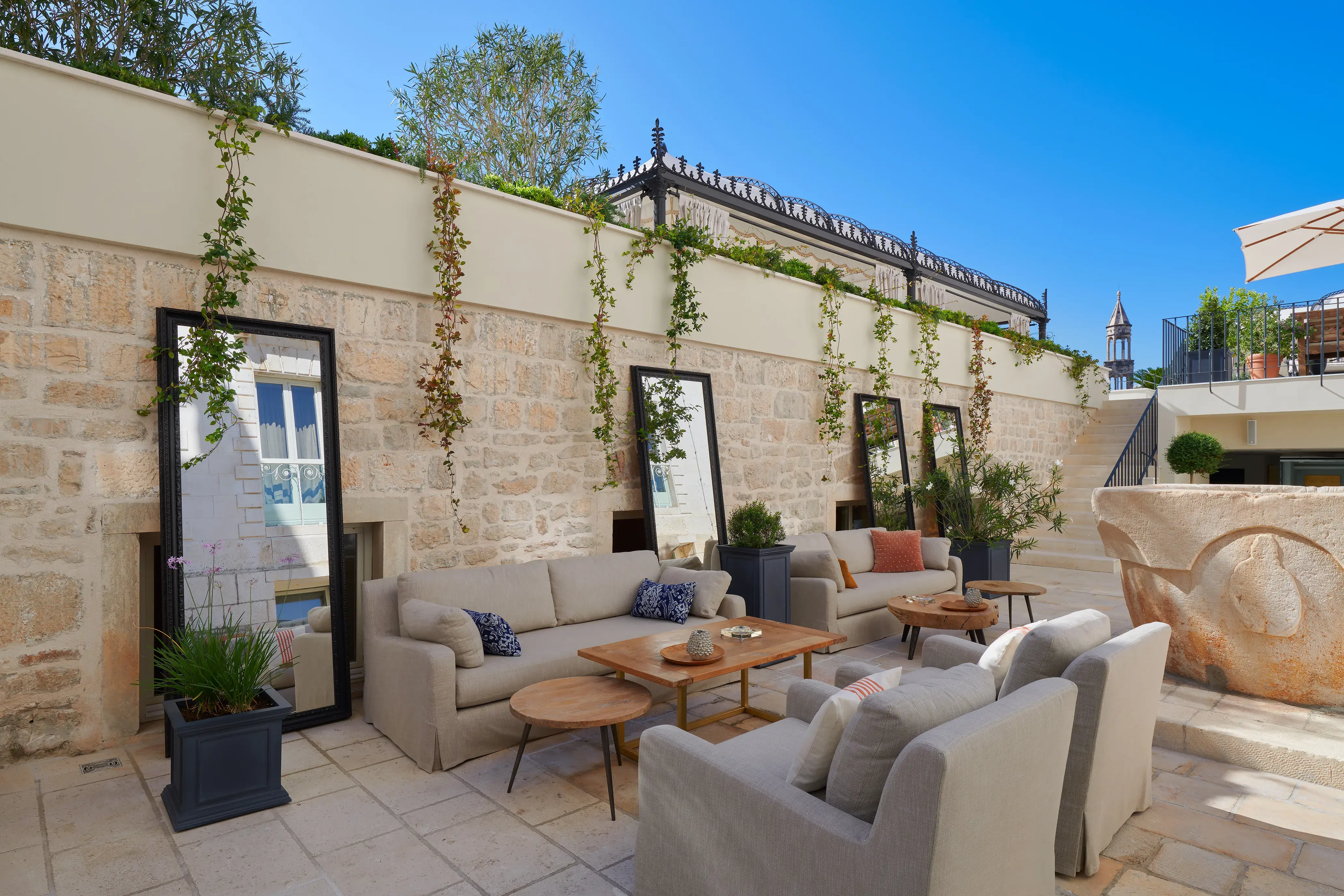 Outdoor seating area at Palace Elisabeth in Croatia featuring comfy sofas and coffee tables set before large mirrors propped up against a stone wall under a clear sky