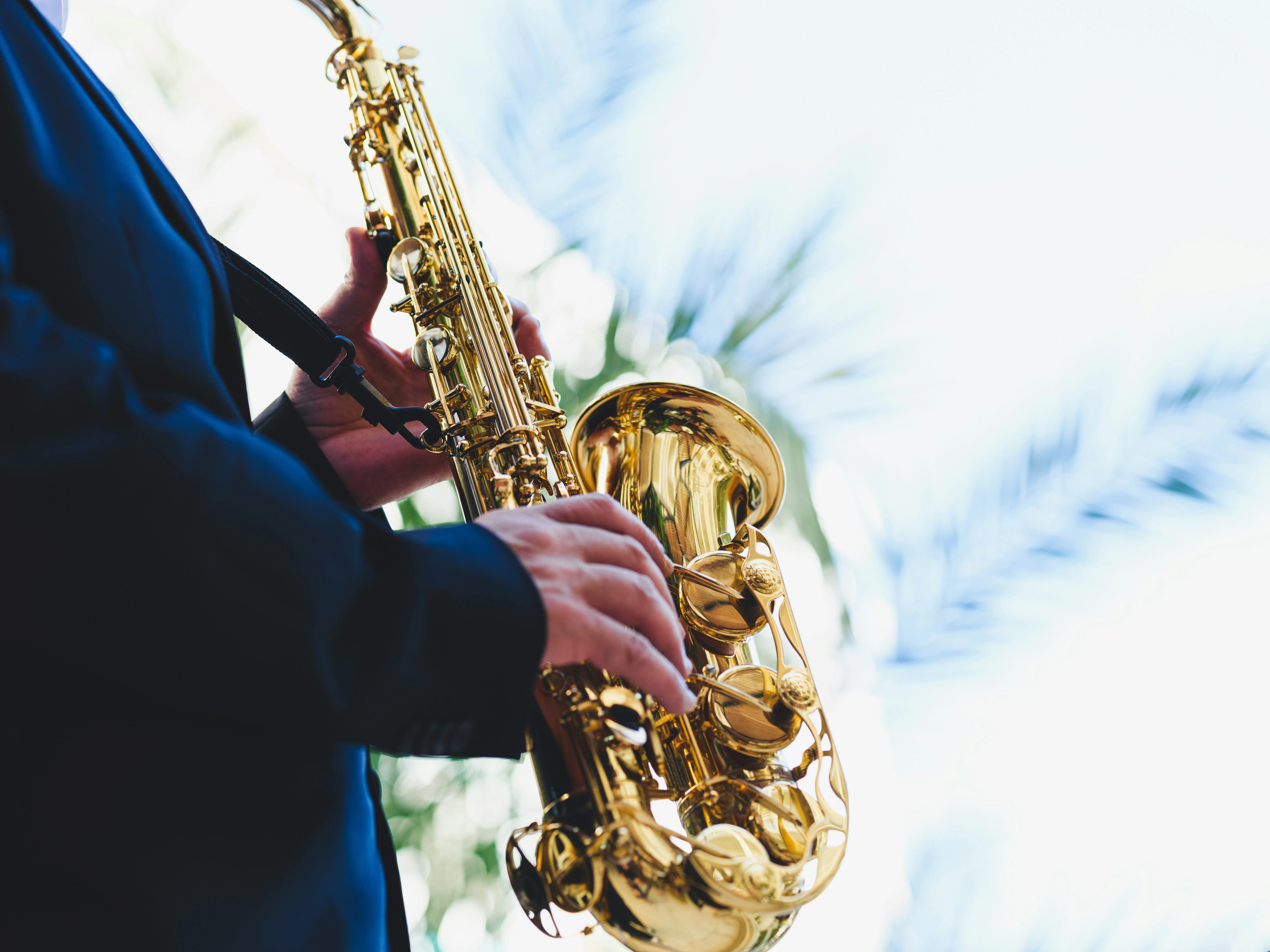 Person playing a shiny gold saxophone outdoors with blurred palm leaves in the background.