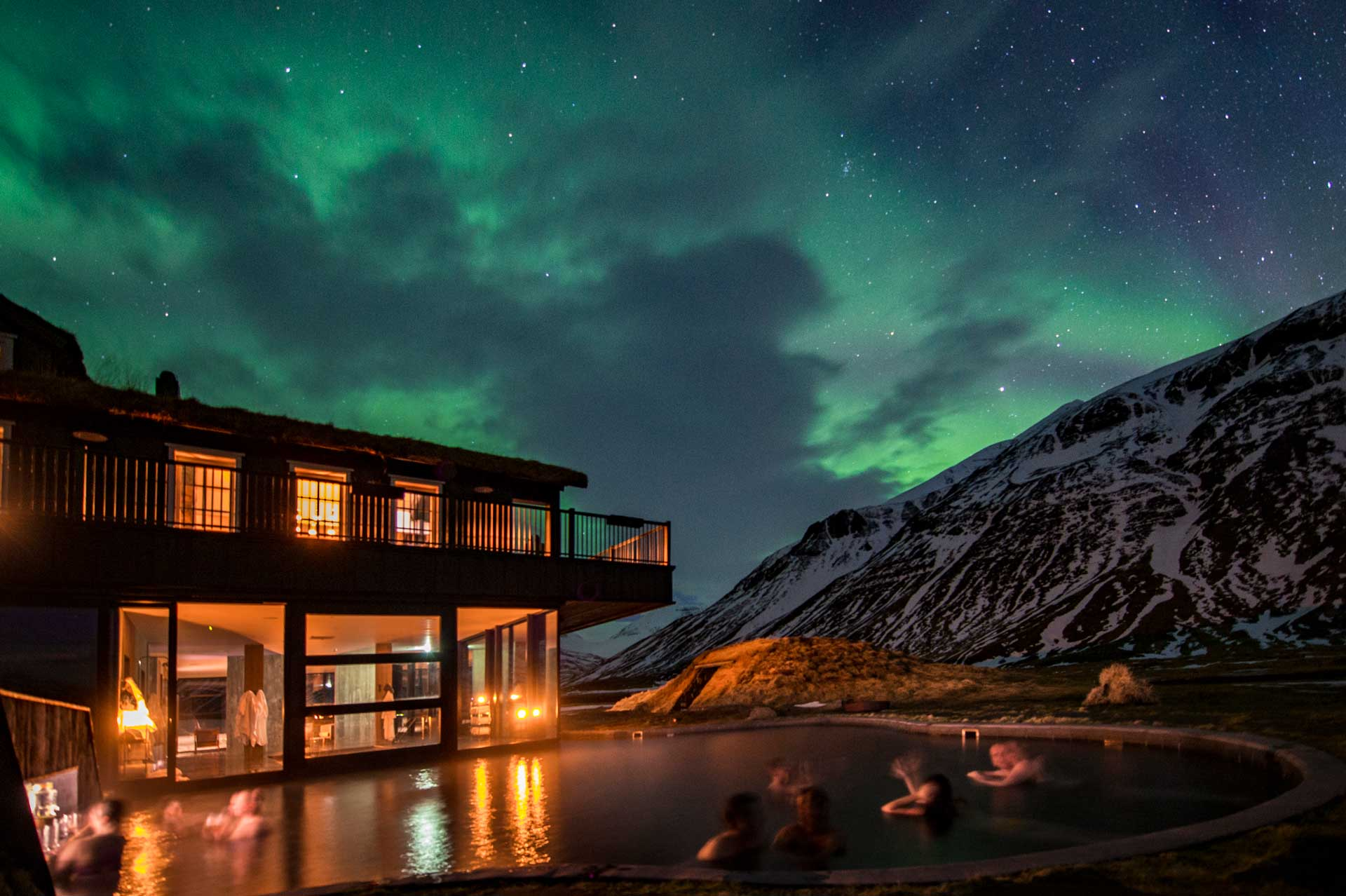 Outdoor geothermal pool at a remote lodge beneath the northern lights, with mountains silhouetted in the background.