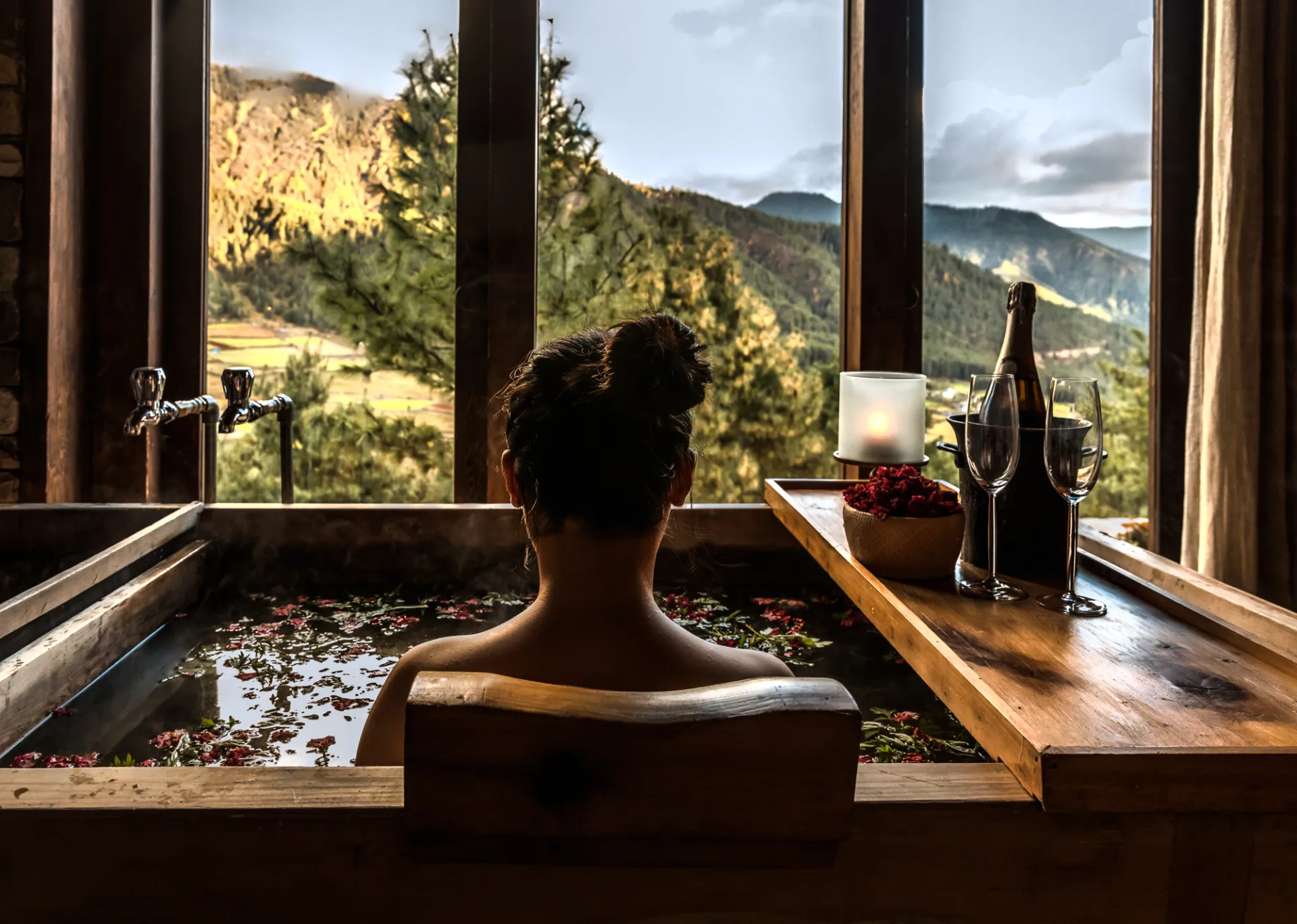 Person relaxing in a flower‑filled bath overlooking forested mountains through large windows.