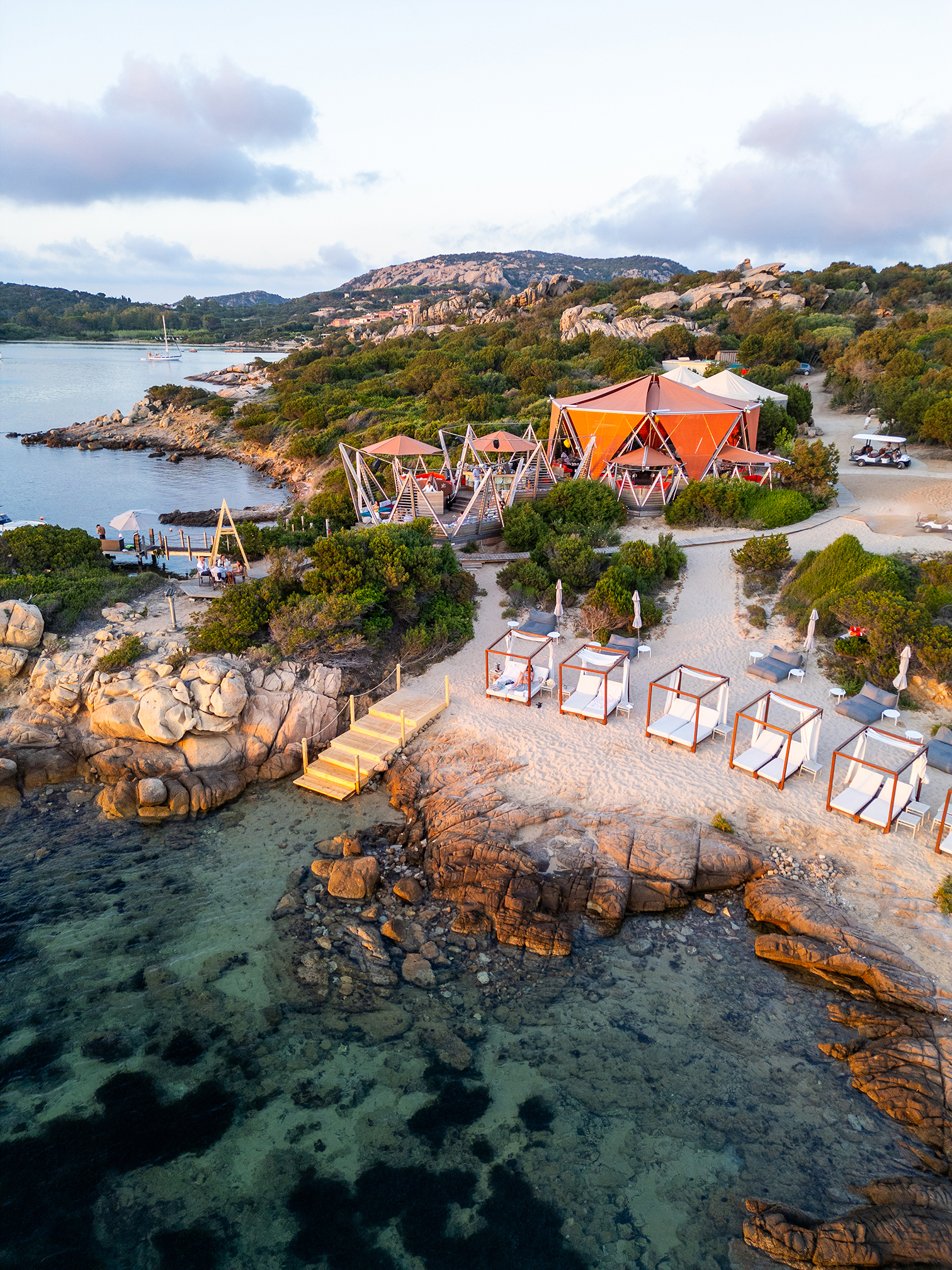 A large orange tented structure with beach cabanas set on a rocky outcrop beside the ocean