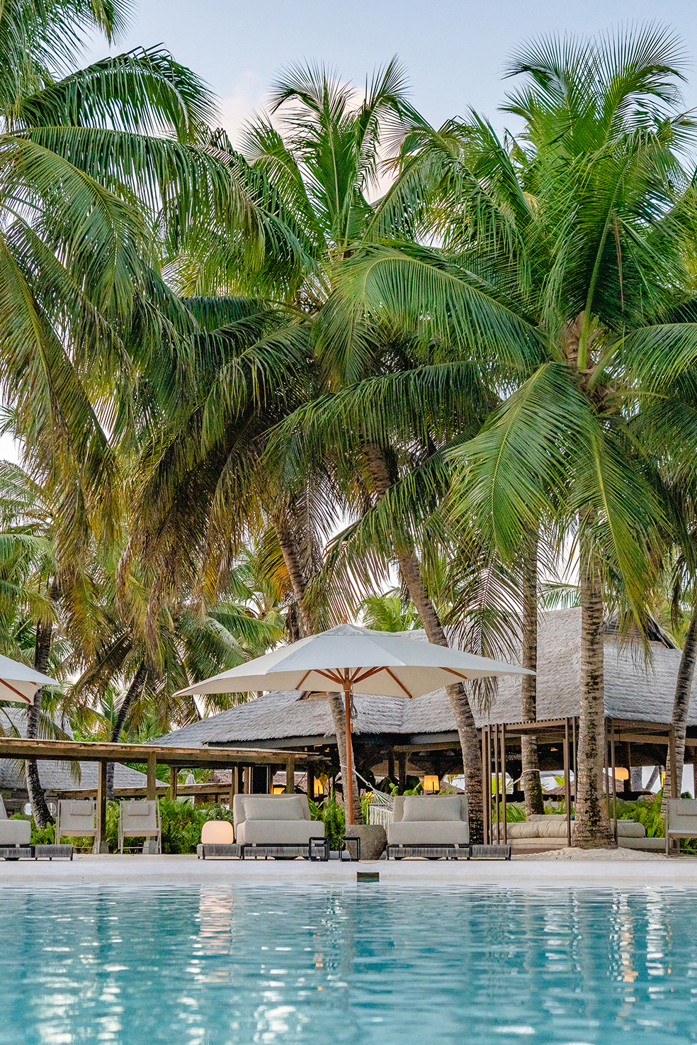 A line of white cabanas and palms beside a tranquil pool at sunset