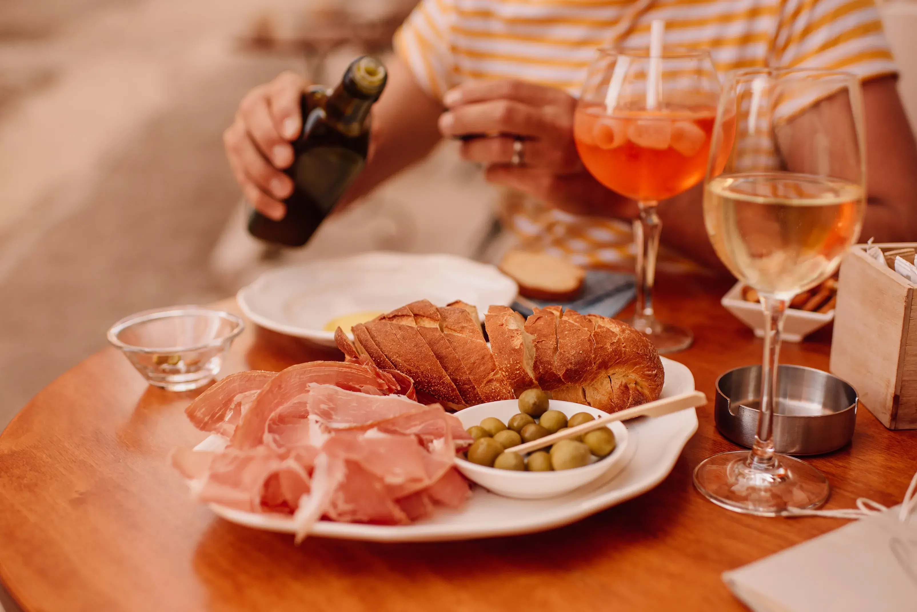 A person ready to pour olive oil to eat a platter of cured meat, bread and olives on a table set next to two glasses of wine