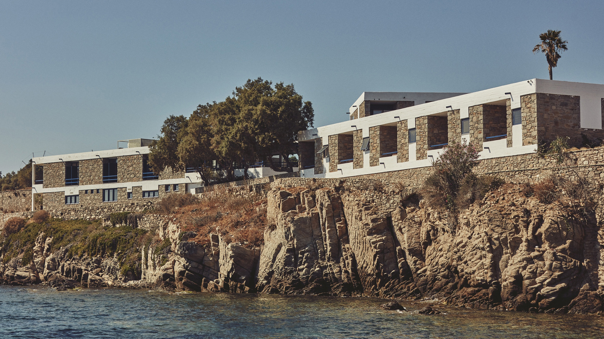 A stone and white building with trees standing on the edge of a rocky outcrop next to the sea