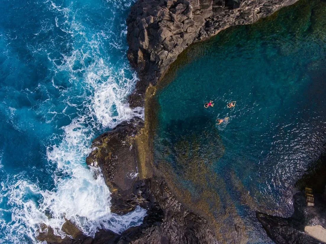 Aerial view of swimmers in a natural ocean rock pool beside crashing waves.
