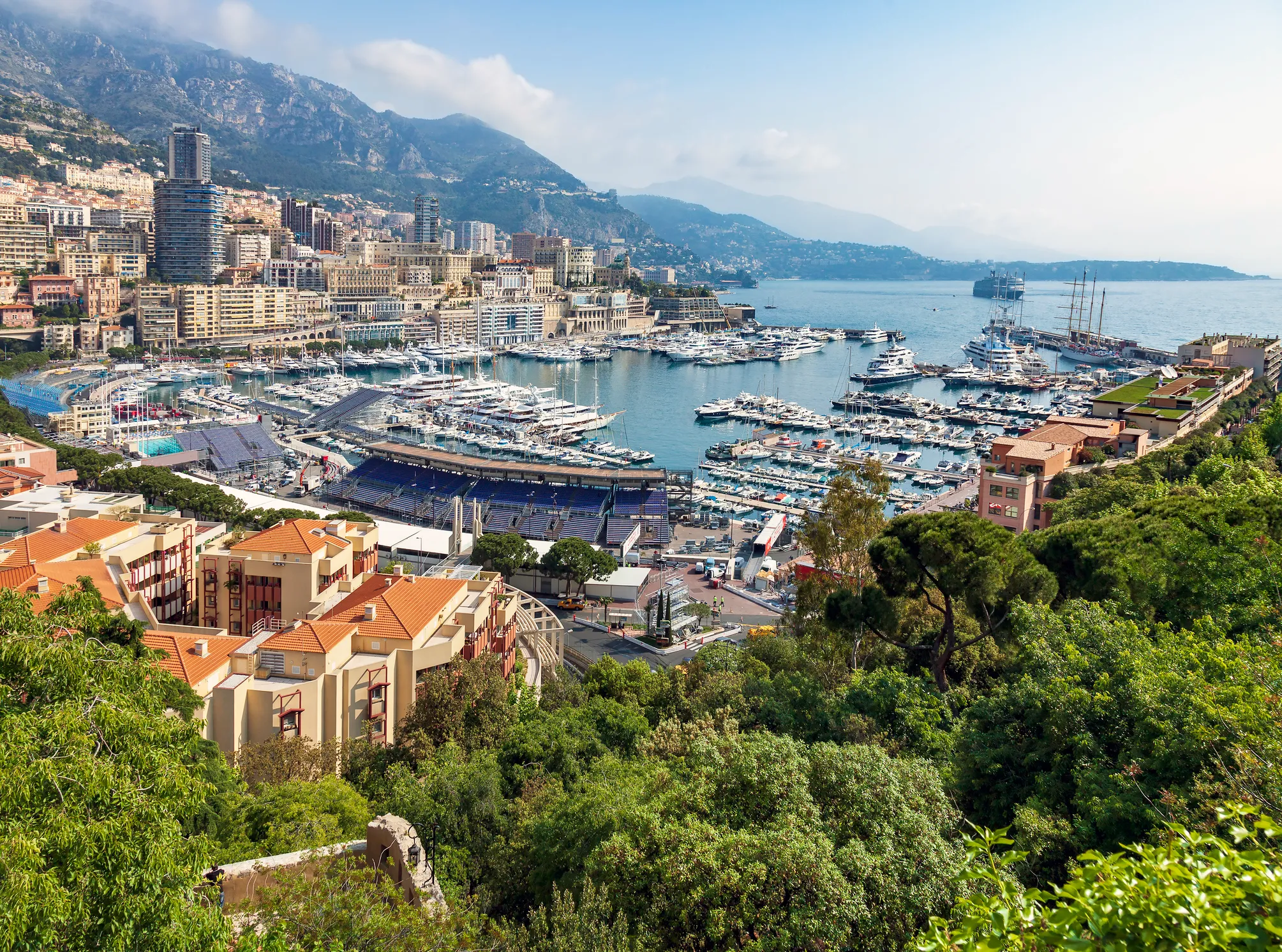 Scenic view of Monaco’s harbor with yachts, surrounded by buildings, greenery, mountains, and the Mediterranean Sea.