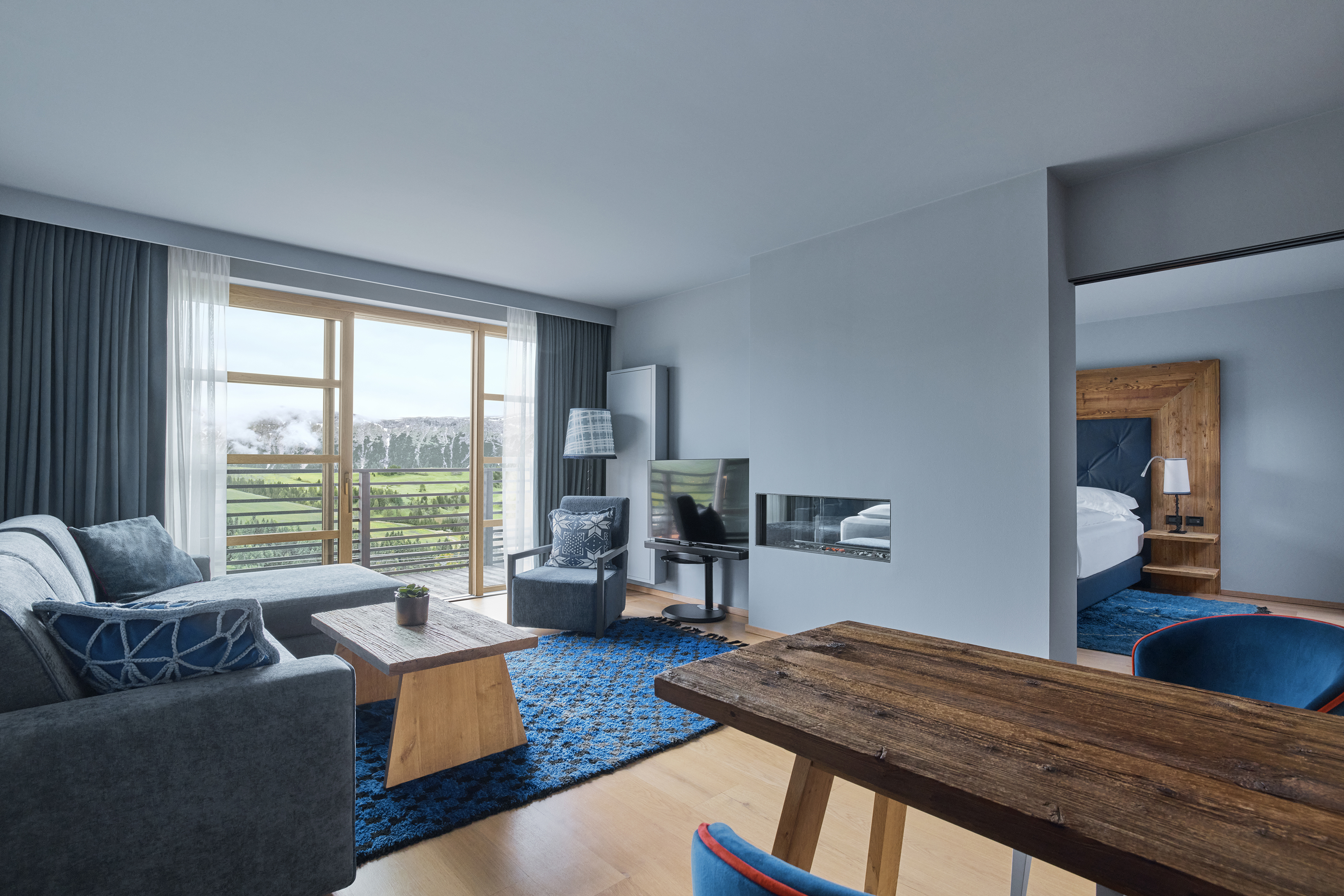 A well-lit bedroom with a large bed, wooden headboard, and a balcony overlooking mountainous scenery.