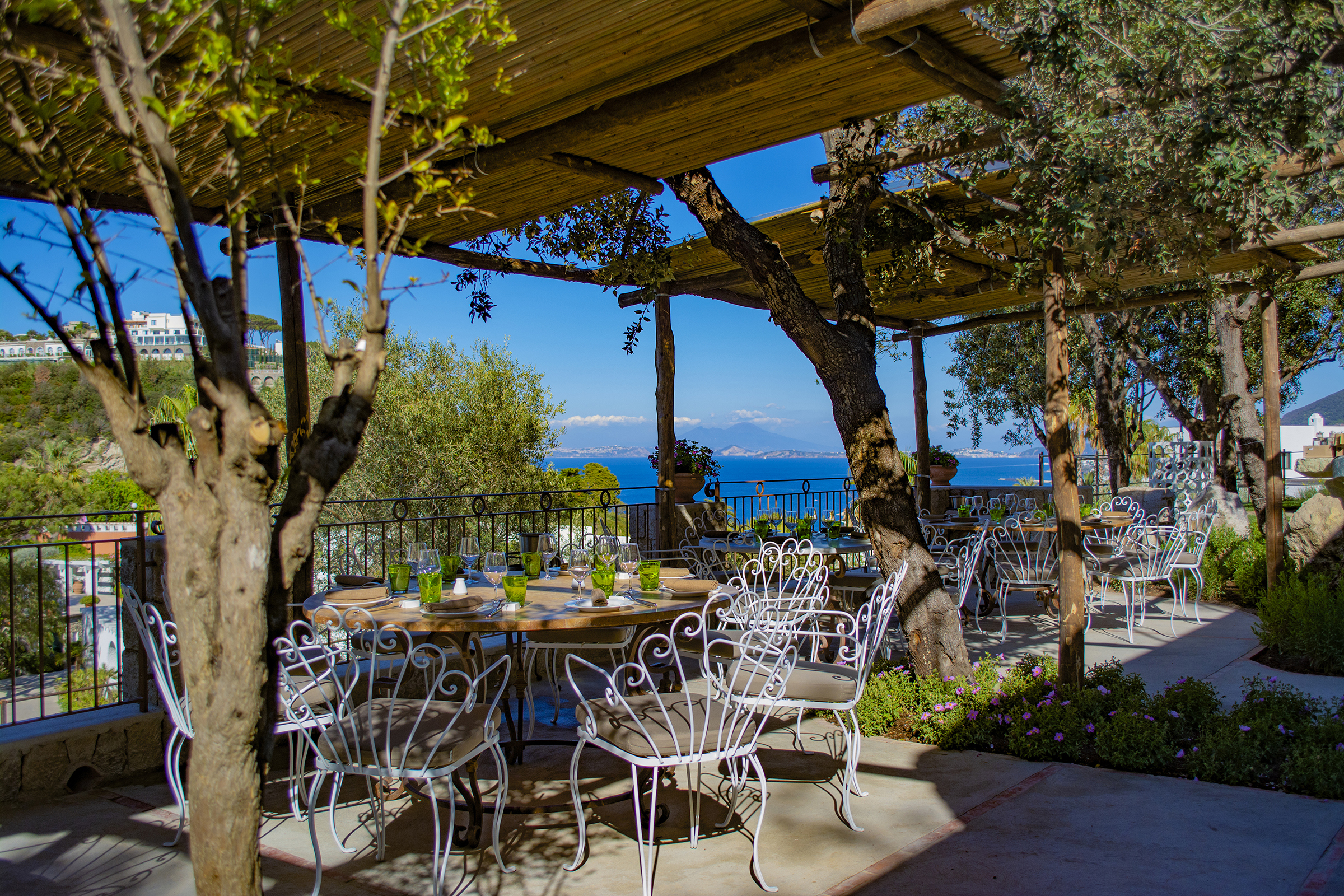 Outdoor dining area of Nonna Marì Farmer Restaurant at Botania Relais & Spa in Ischia with elegant white wrought iron furniture, shaded by a wooden pergola, overlooking lush greenery and the sea.