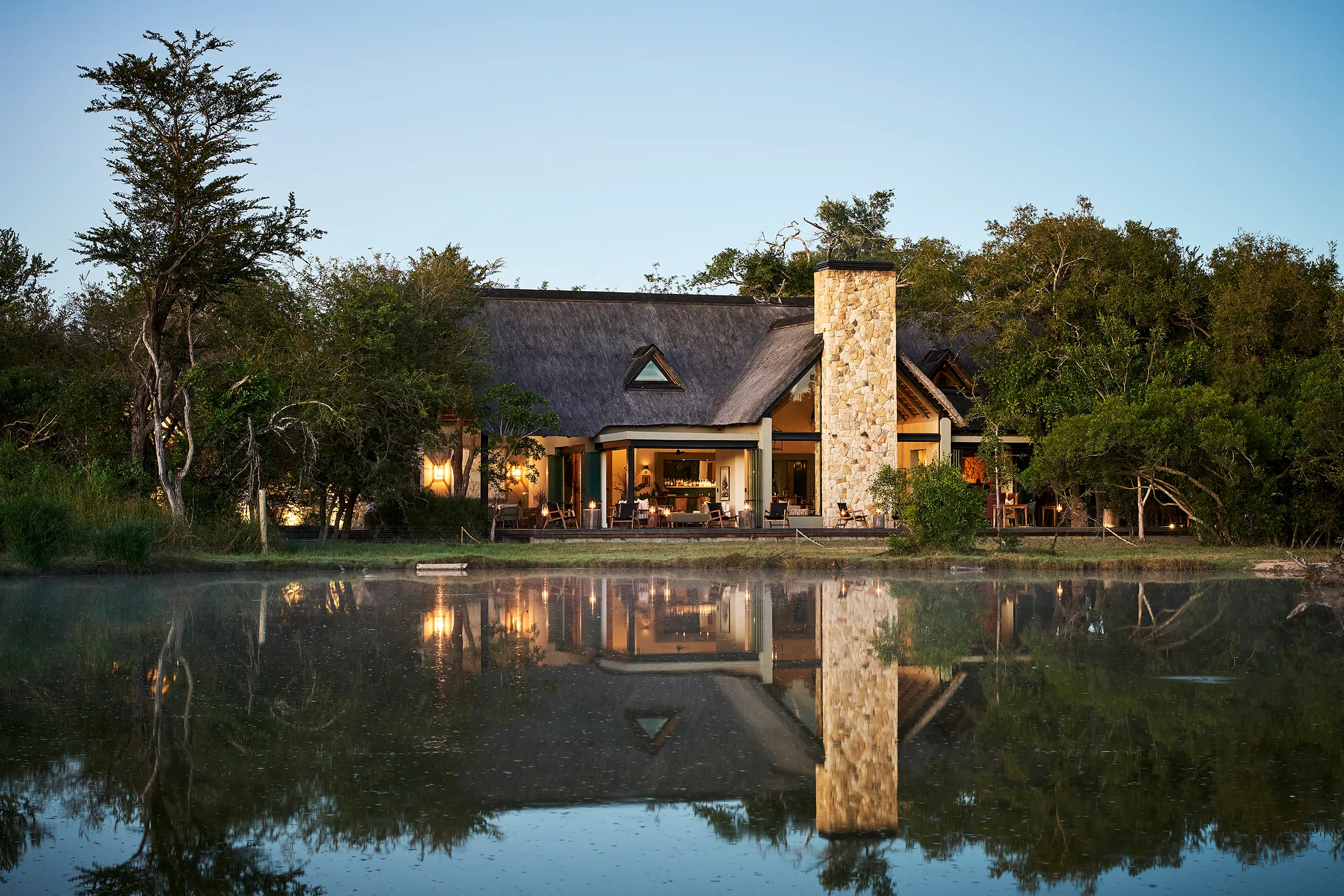 The thatched and stone exterior of Monwana Game Reserve featuring a large chimney surrounded by tall trees from across a tranquil lake 