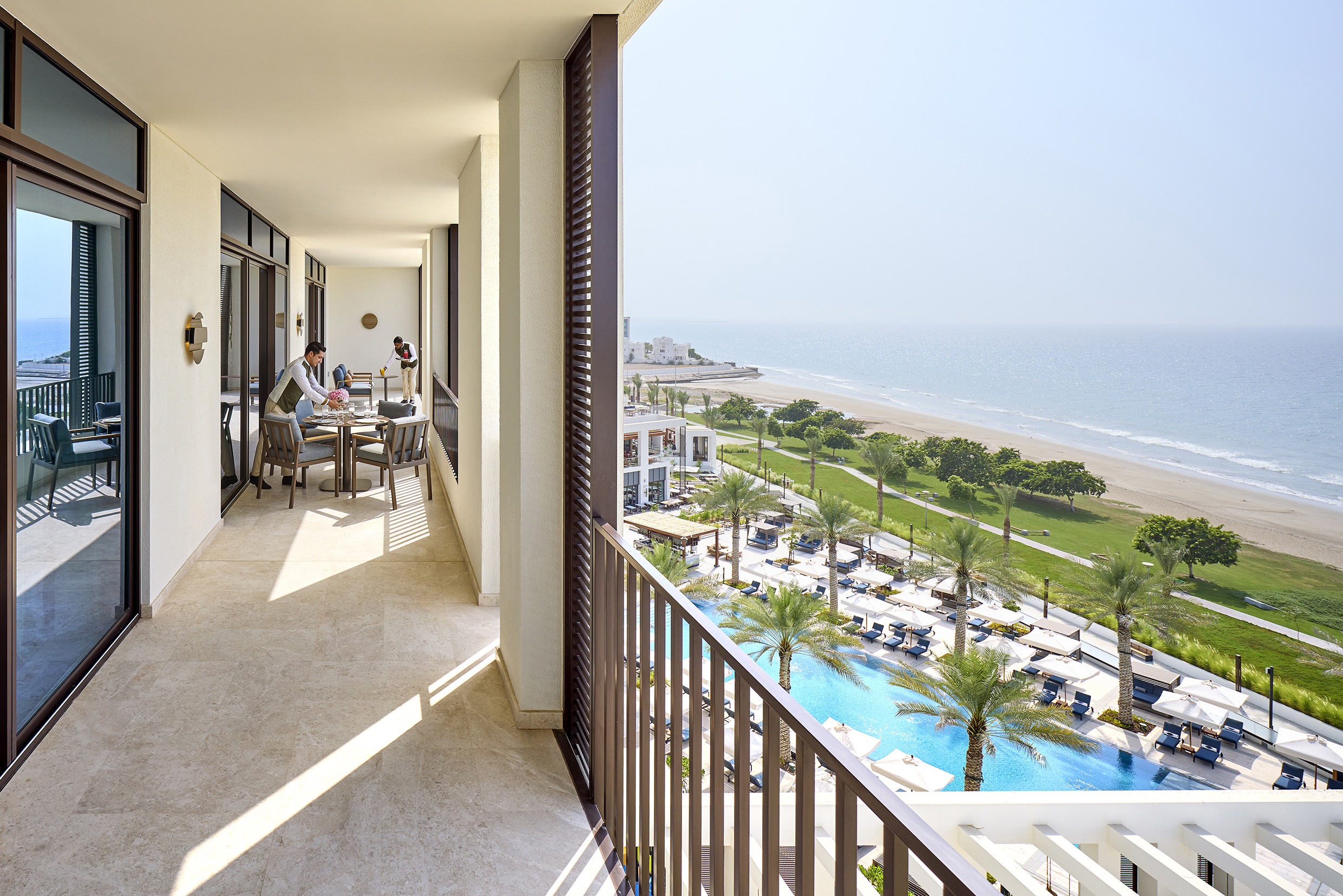 The long terrace of a room at Mandarin Oriental Muscat with a butler setting a table for food, featuring sweeping view of the beach and pool in front of the hotel