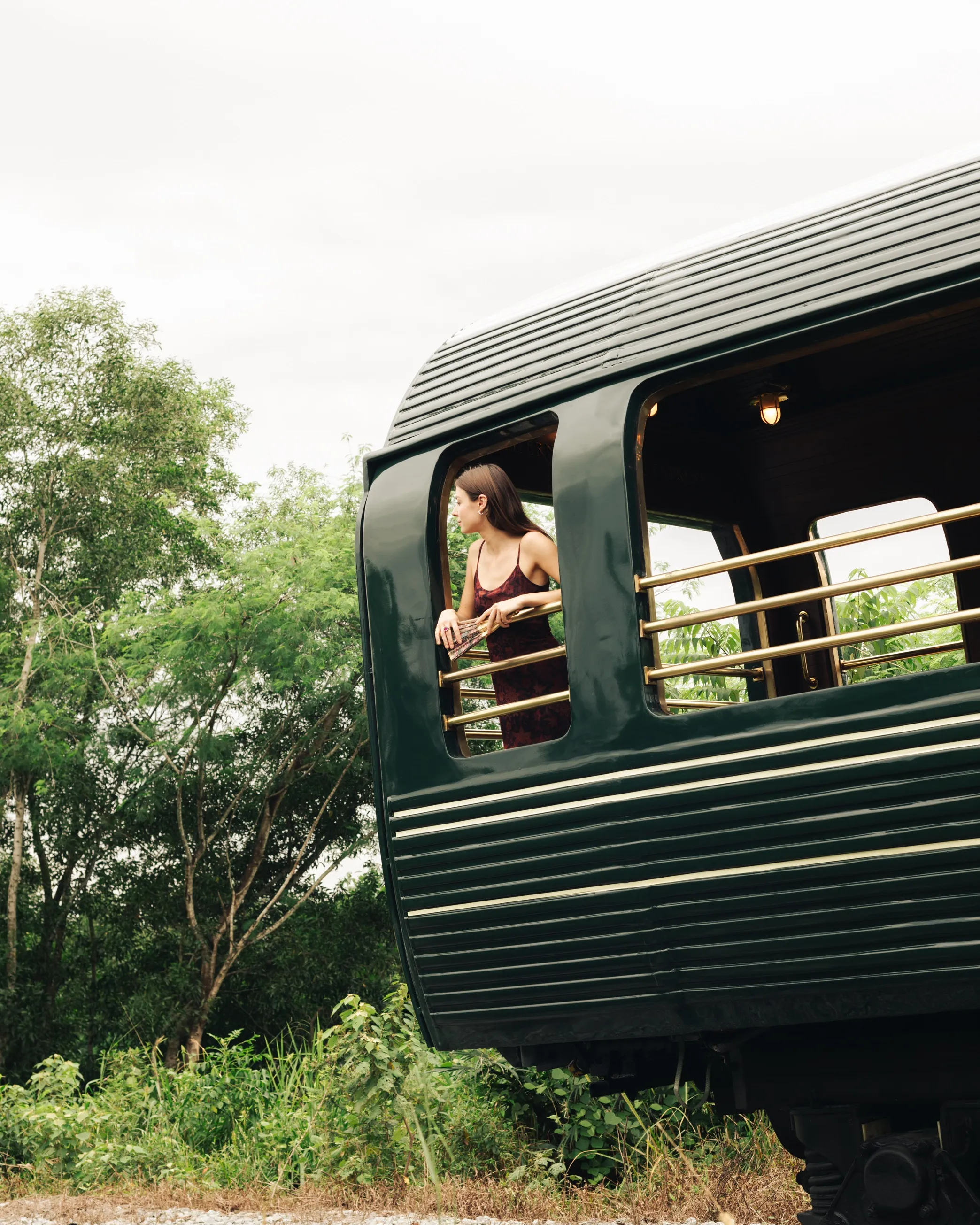 A woman stands on the balcony of a green train car, looking out at the surrounding greenery.