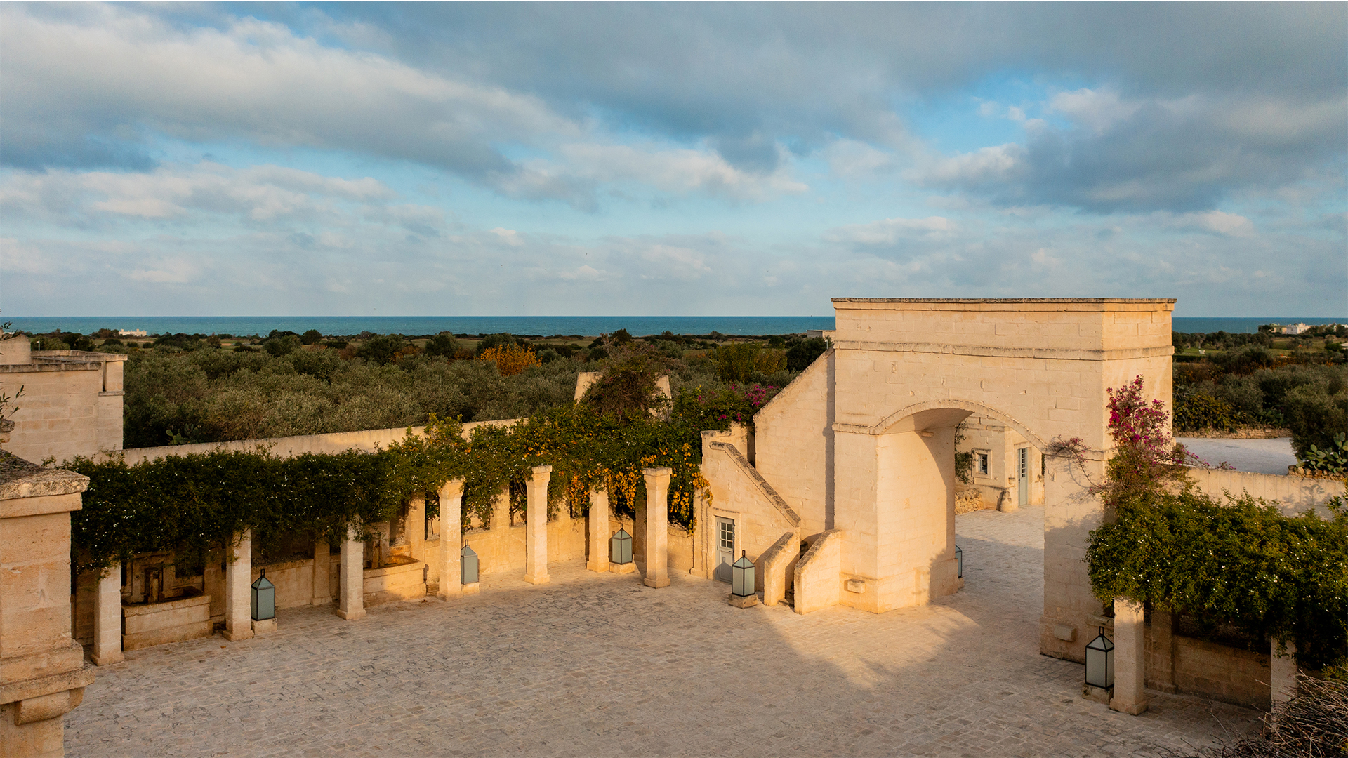 A tranquil courtyard with an arched entrance, surrounded by greenery under a clear sky at dusk.