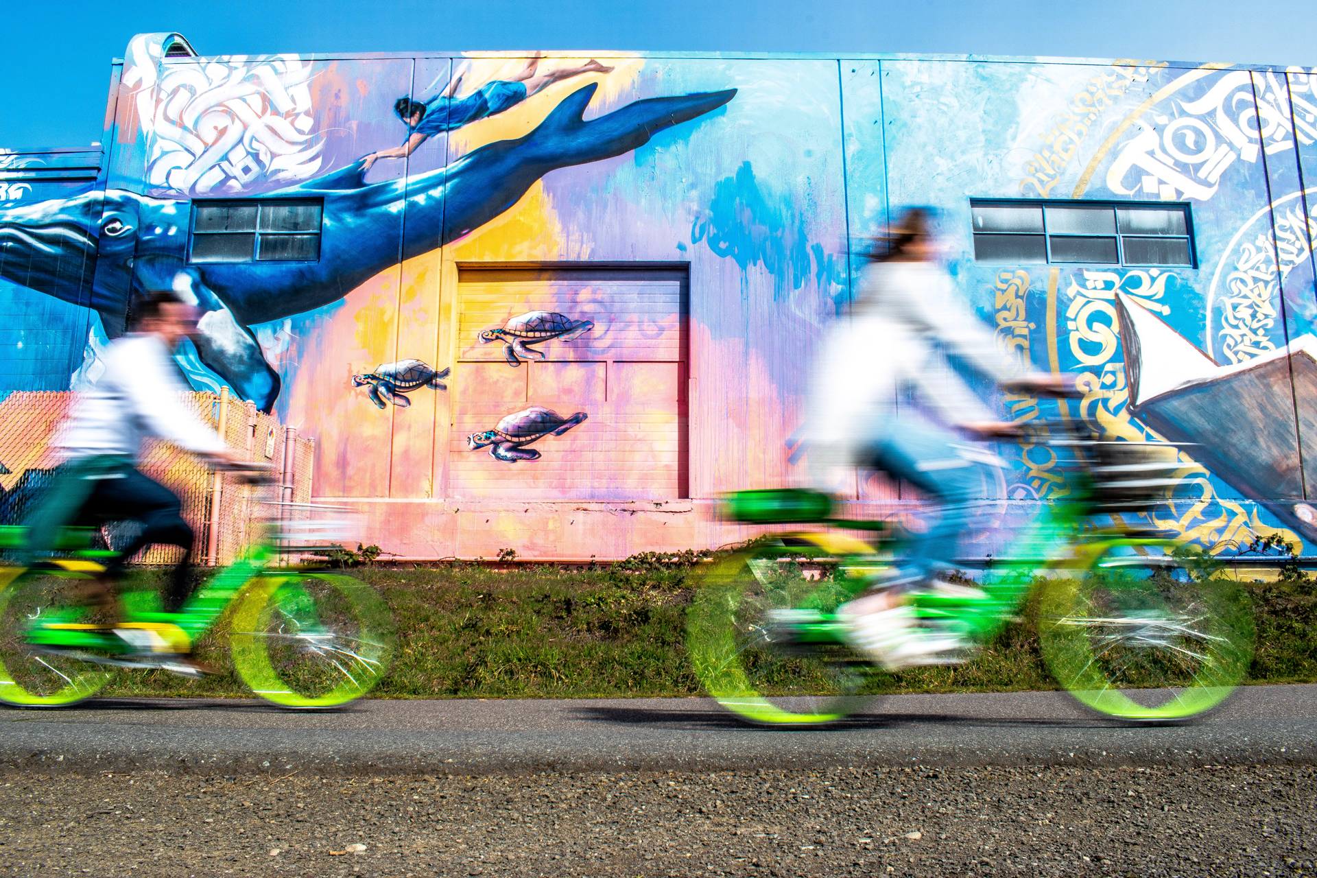Two people blurred on green bicyles in front of street art