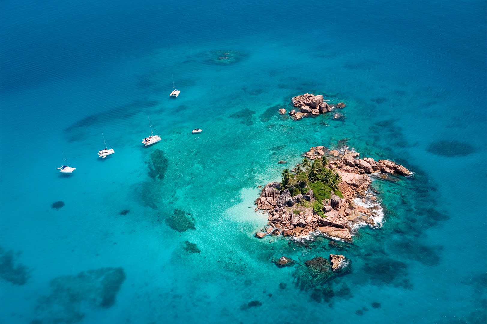 Aerial view of a small, rocky island surrounded by clear turquoise waters with several boats anchored nearby.