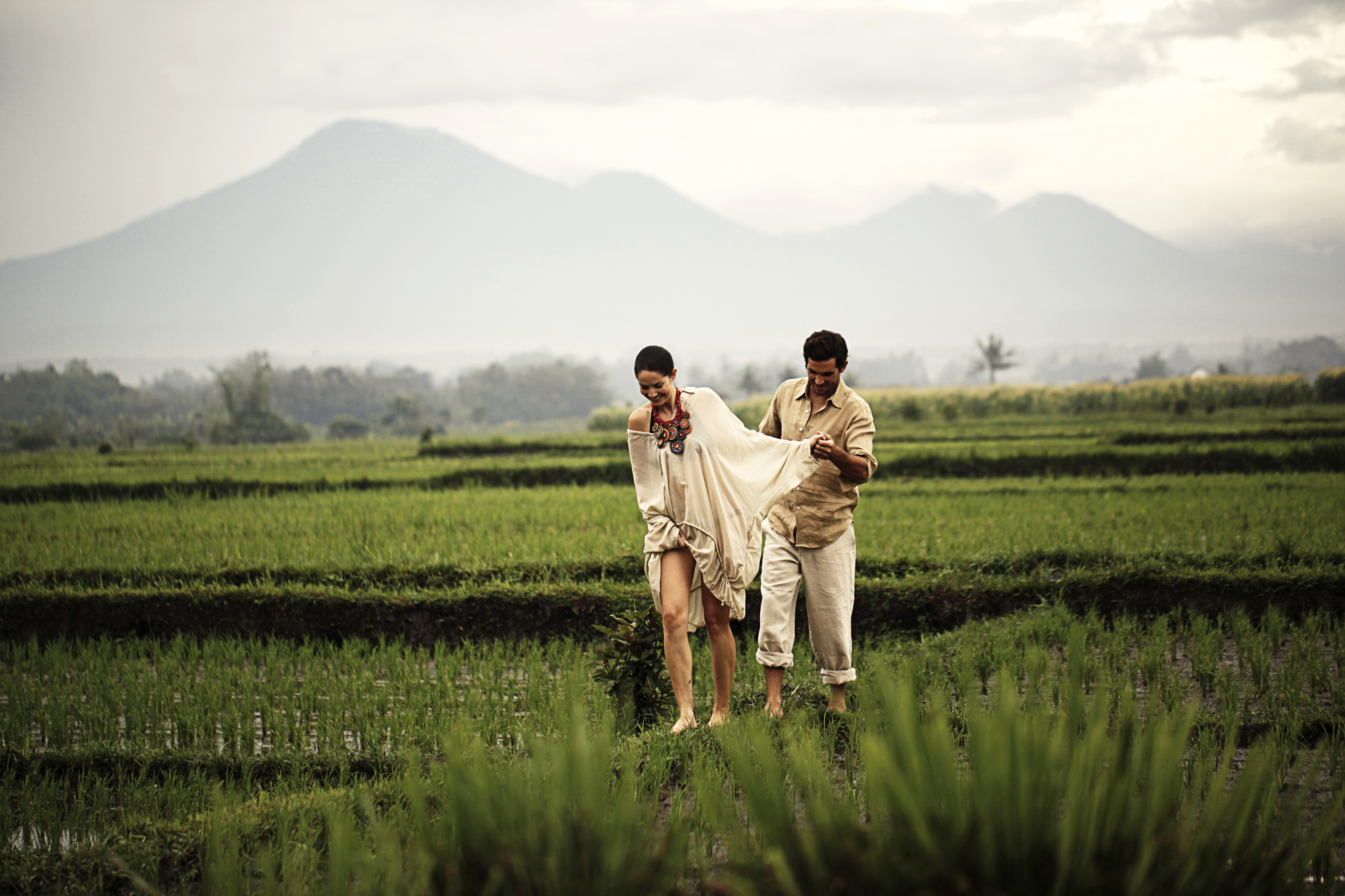 A couple walks through a field of green crops with misty mountains seen in the background