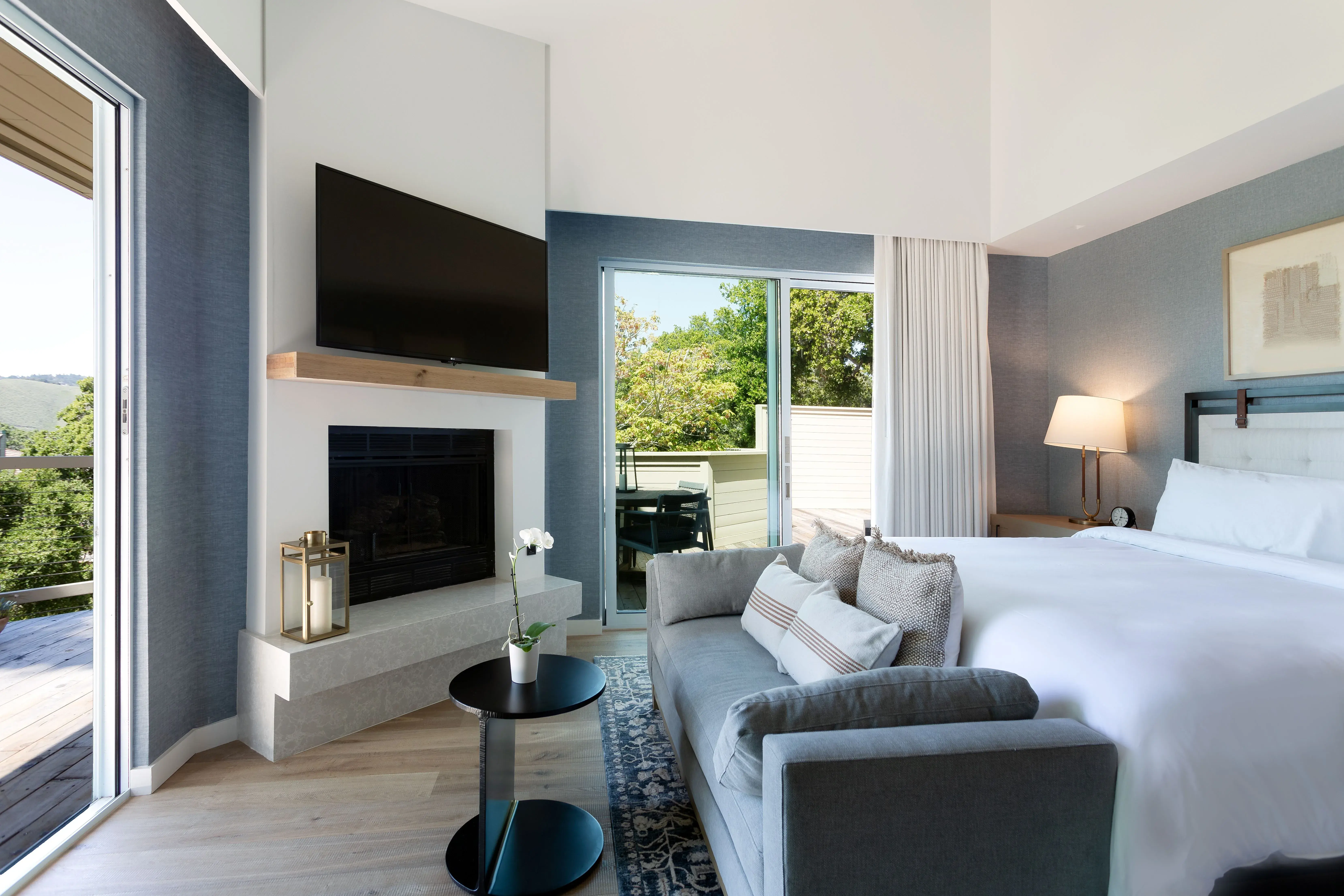 A sleek Family Suite at Carmel Valley Ranch in California featuring a plush bed with a sofa in front, a fireplace and tv with two large doors showing panoramic views