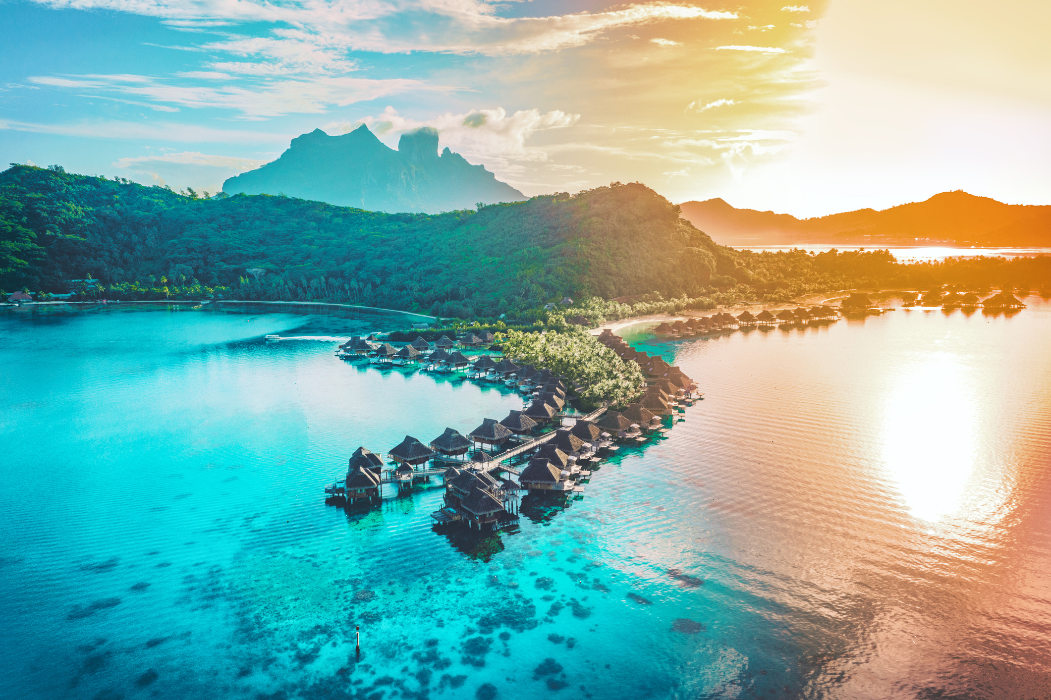 Aerial view of an overwater bungalow resort with a mountain in the background at sunset.