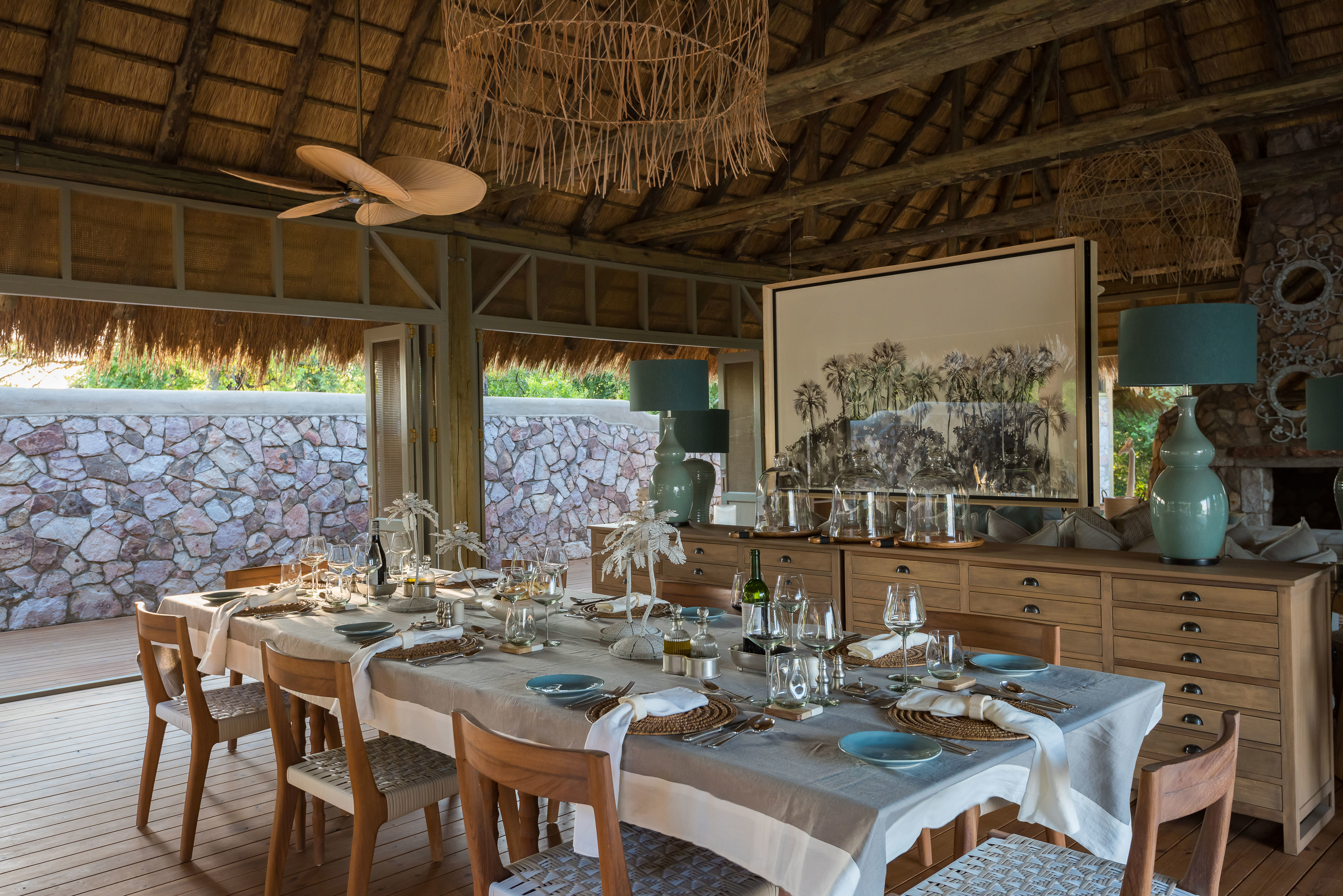 A large dining table set for a meal inside a thatched building beside a large chest of drawers and artwork