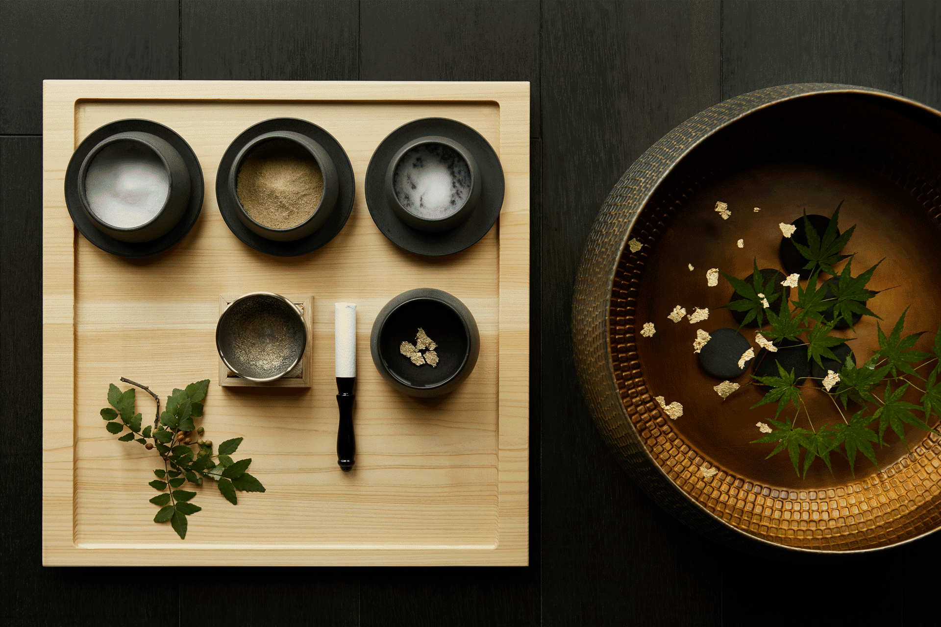 Traditional spa ritual setting with bowls of natural ingredients arranged beside a copper basin filled with floating leaves and petals.