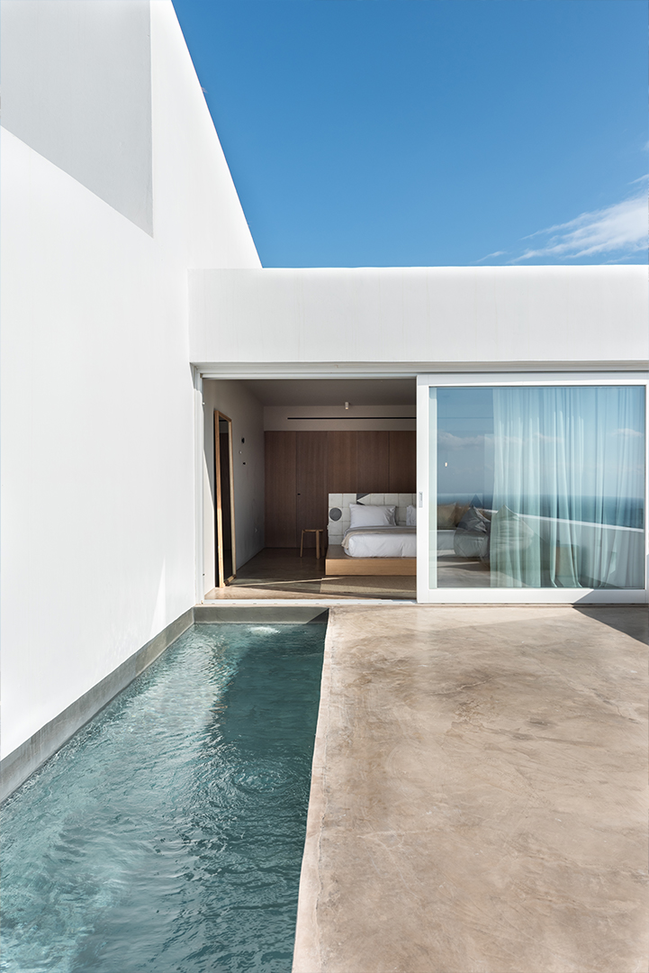 Overlooking a whitewashed villa, with a view of a private plunge pool and looking into its open patio door where a plush white bed and wood toned furniture are seen