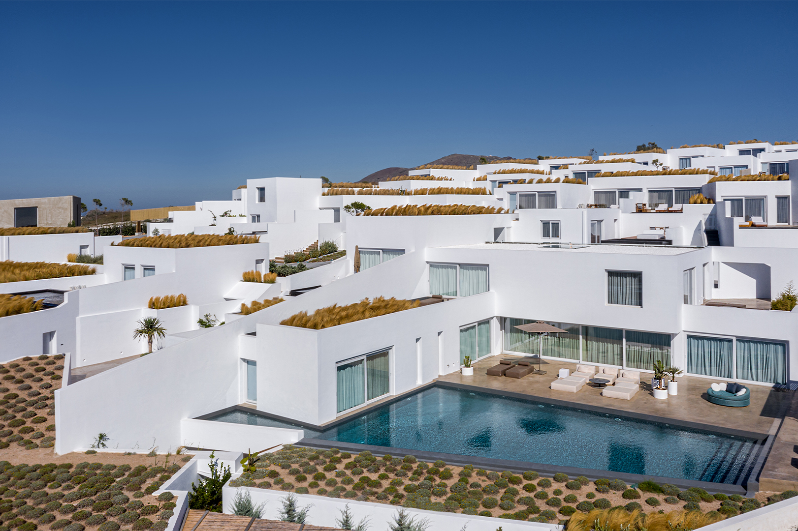 A series of whitewashed buildings and a plunge pool are surrounded by a Greeky grassy landscape
