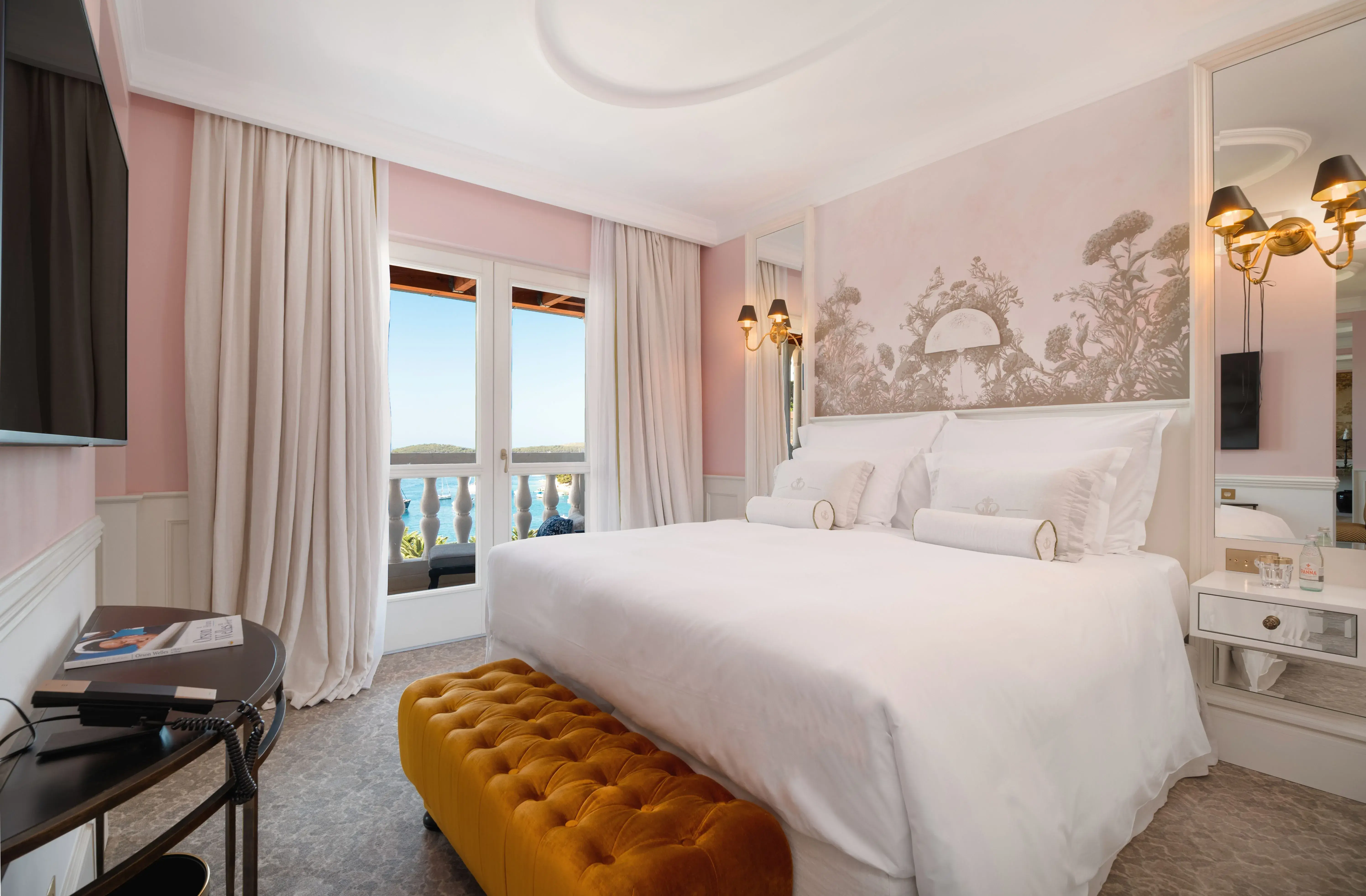 Bedroom of the Presidential Penthouse Suite at Palace Elisabeth in Croatia featuring a bed, with white linens set before a decorative mural with a gold ottoman, mirrors and pink walls