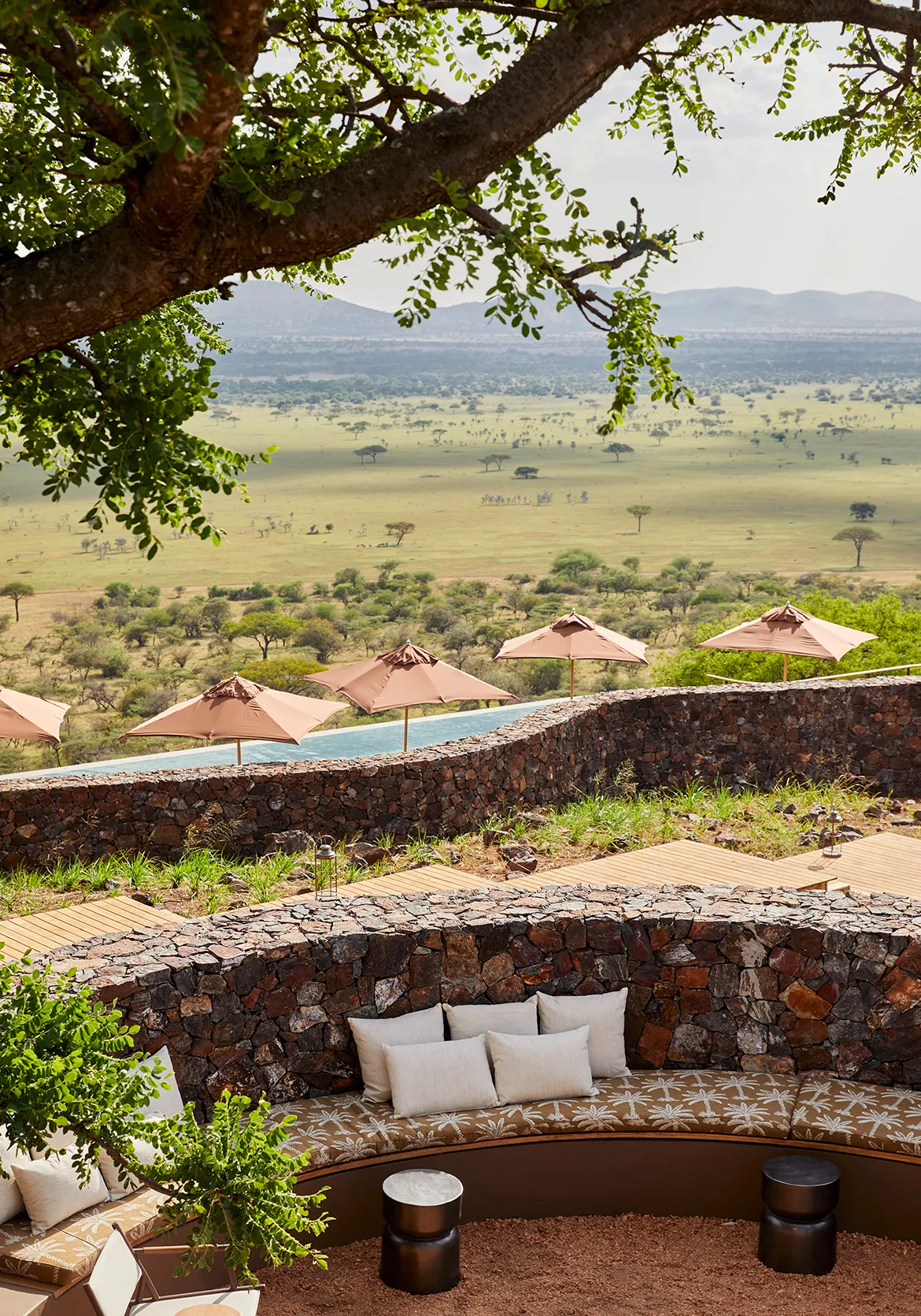 Curved outdoor lounge with cushioned seating overlooking a savannah landscape, framed by stone walls and shaded by trees.