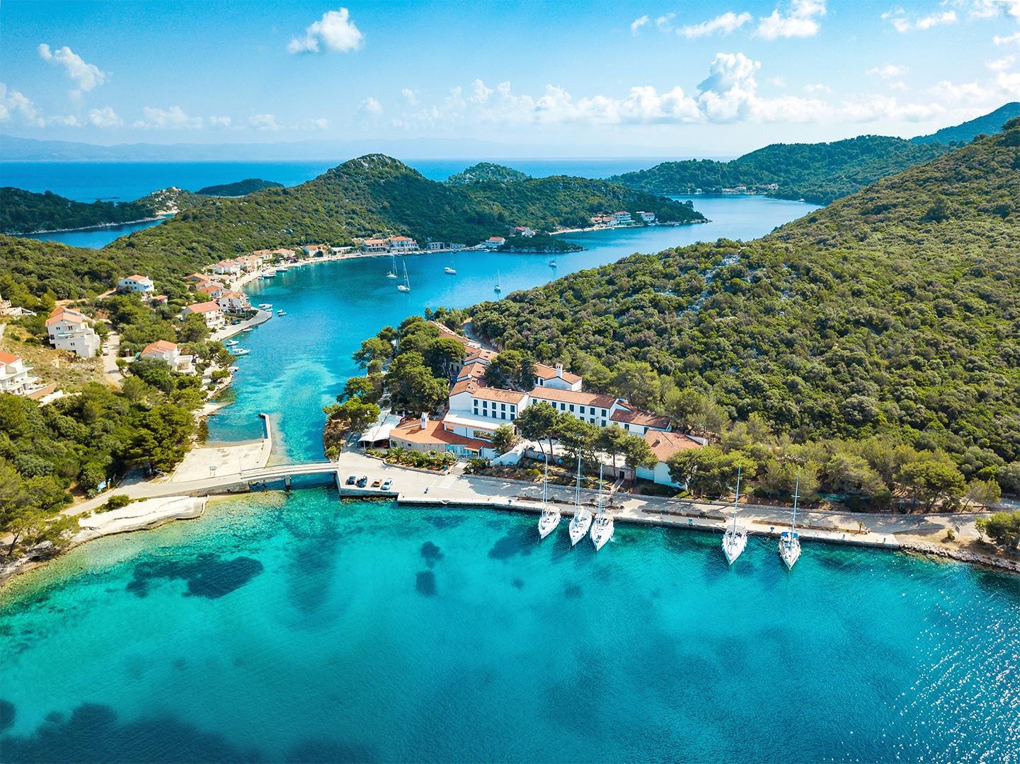 Aerial view of a coastal village with boats docked in a serene blue bay surrounded by lush greenery.
