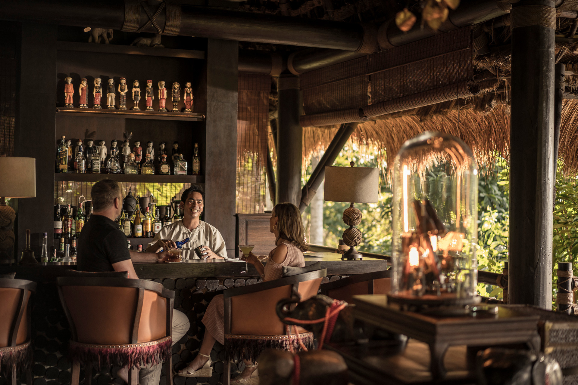 A couple drinking at a rustic chic bar with statues on the wall and a smiling bar tender pouring a drink at Four Seasons Tented Camp