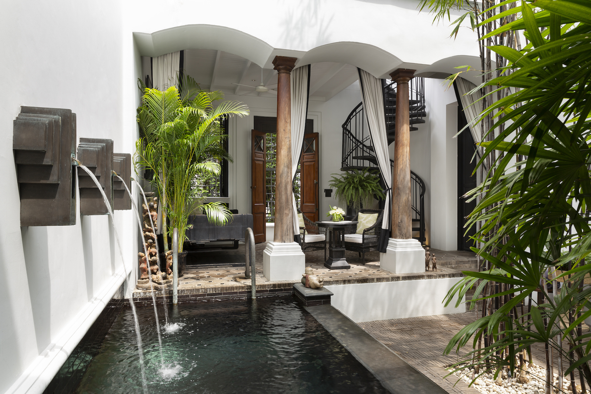 A white courtyard with fountain, pillars and plants featuring a table and chairs and spiral staircase further back at The Siam