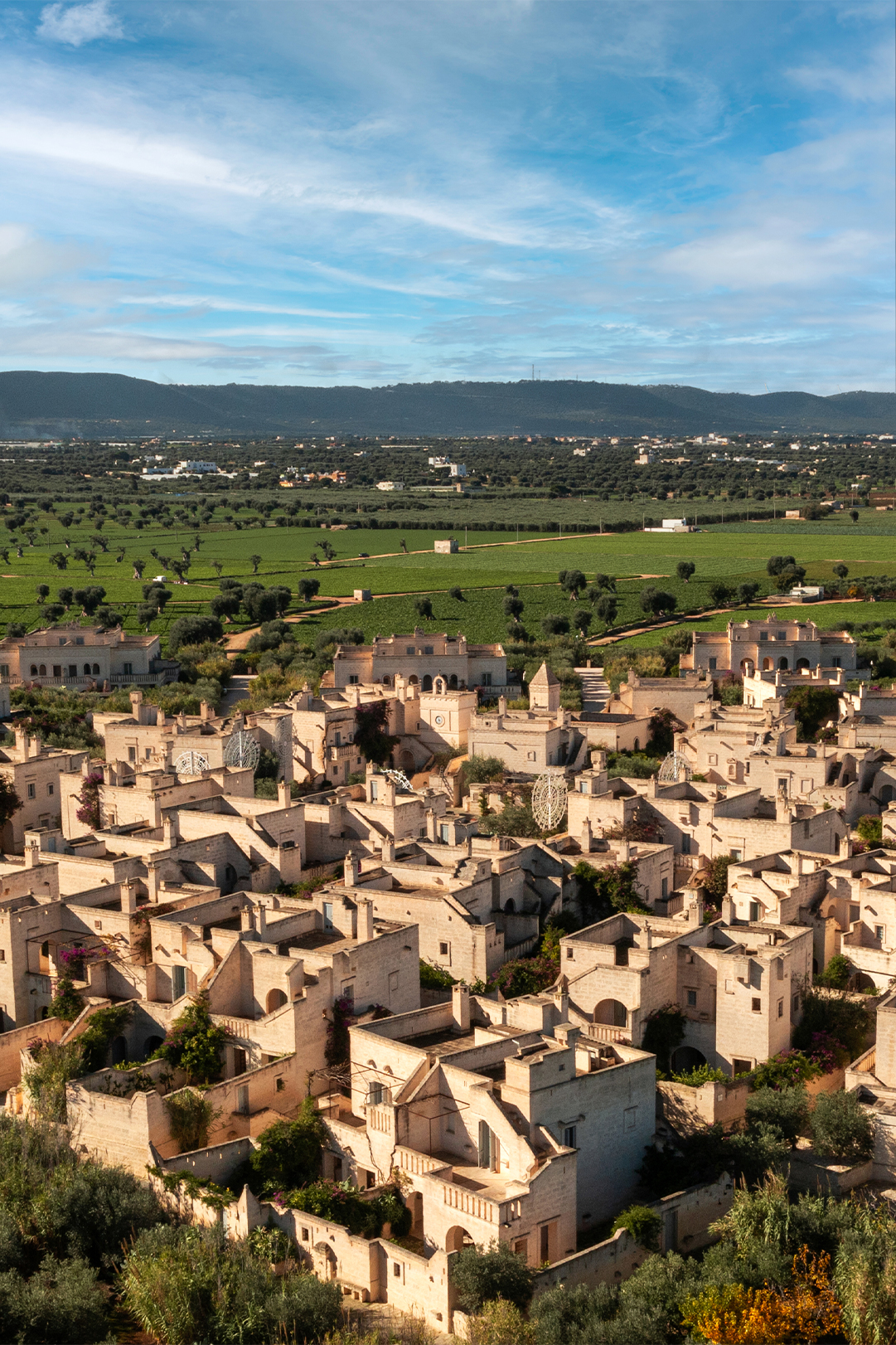 Aerial view of a historic town with dense stone buildings and narrow streets, surrounded by lush greenery and hills under a clear blue sky.