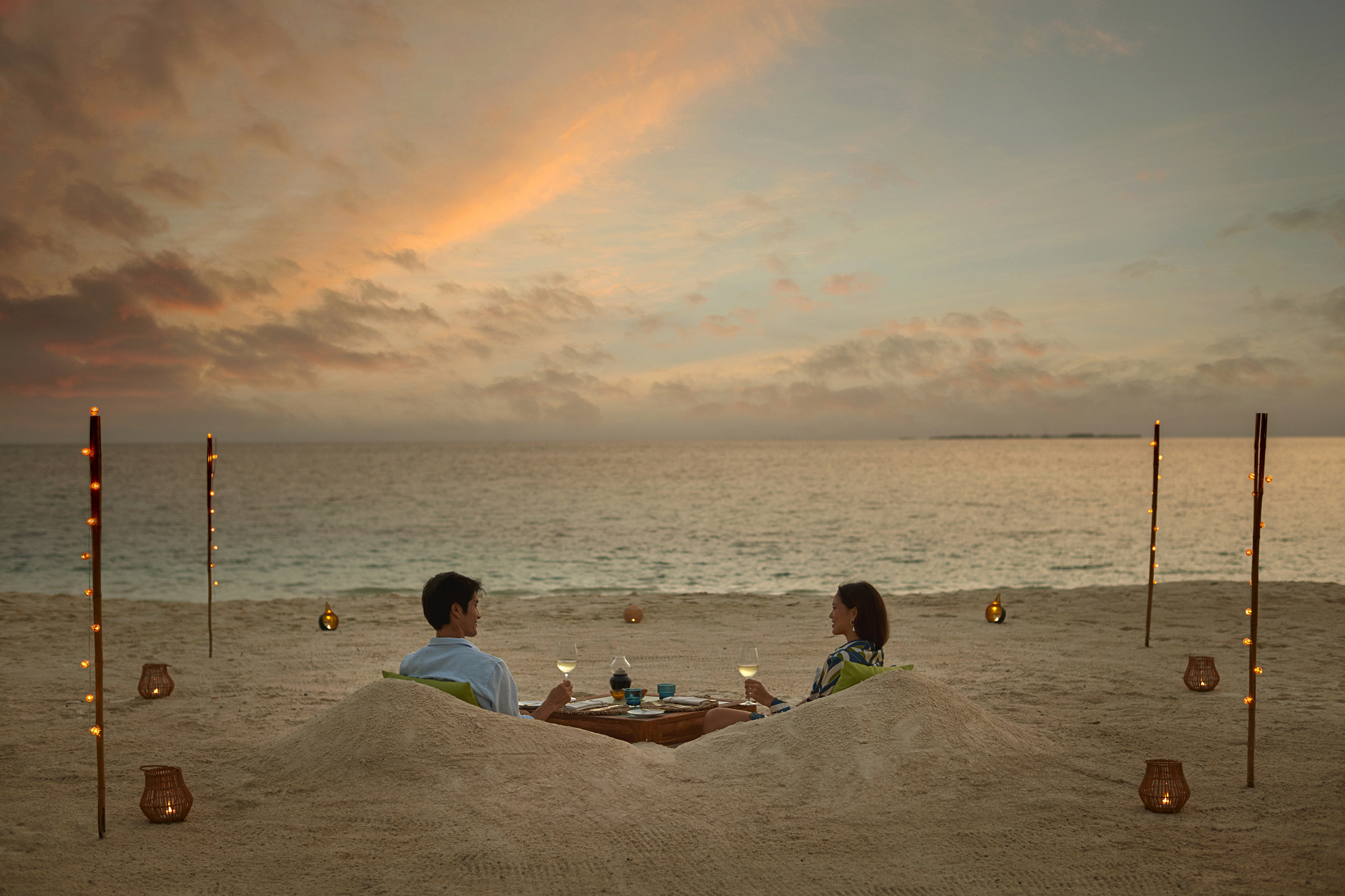 Couple on a private dinner experience in the sand surrouneded by tourches at sunset at Six Senses Kanuhura