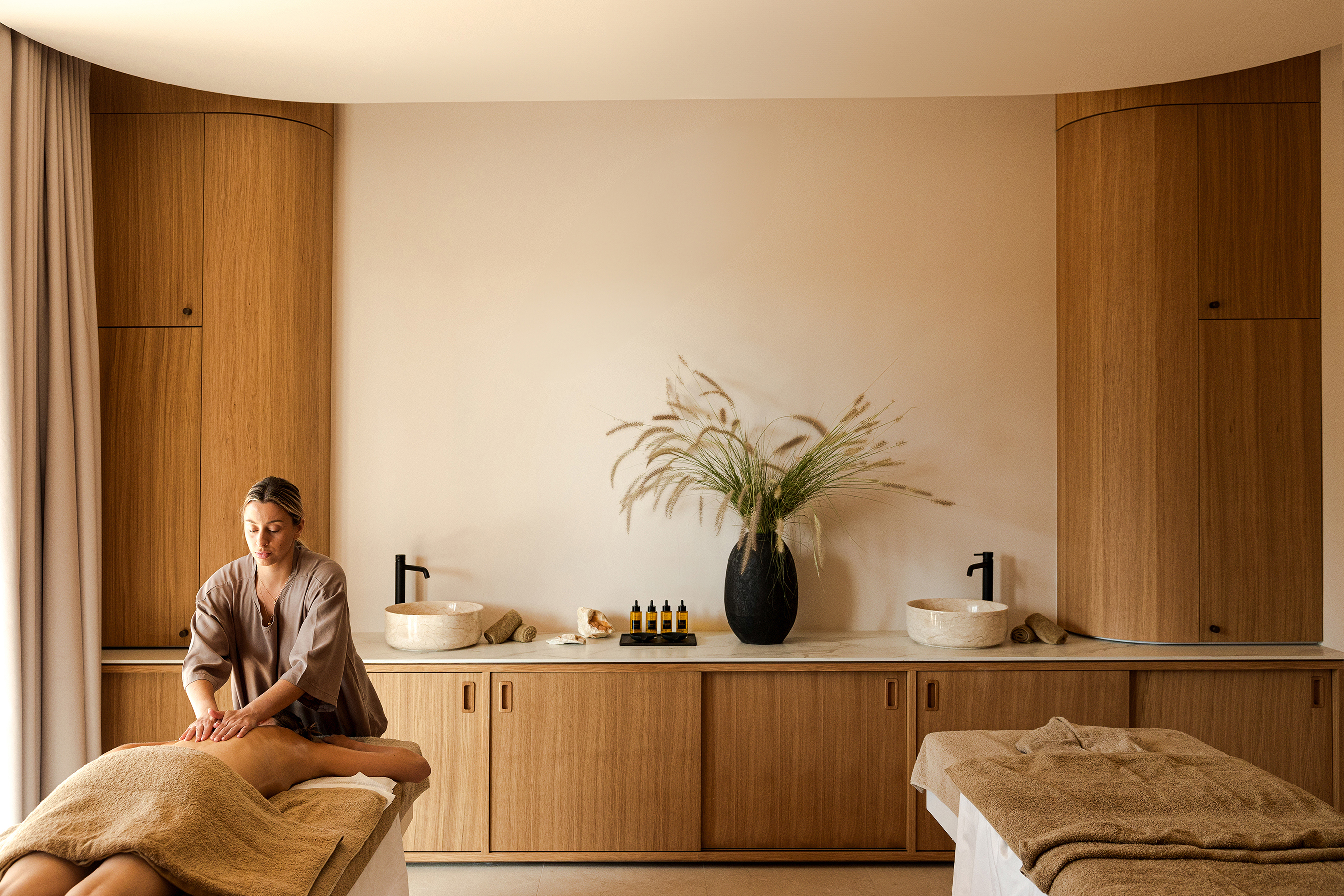 A woman during a massage treatment and a masseuse in a large relaxing spa room featuring two beds and sleek furnishings
