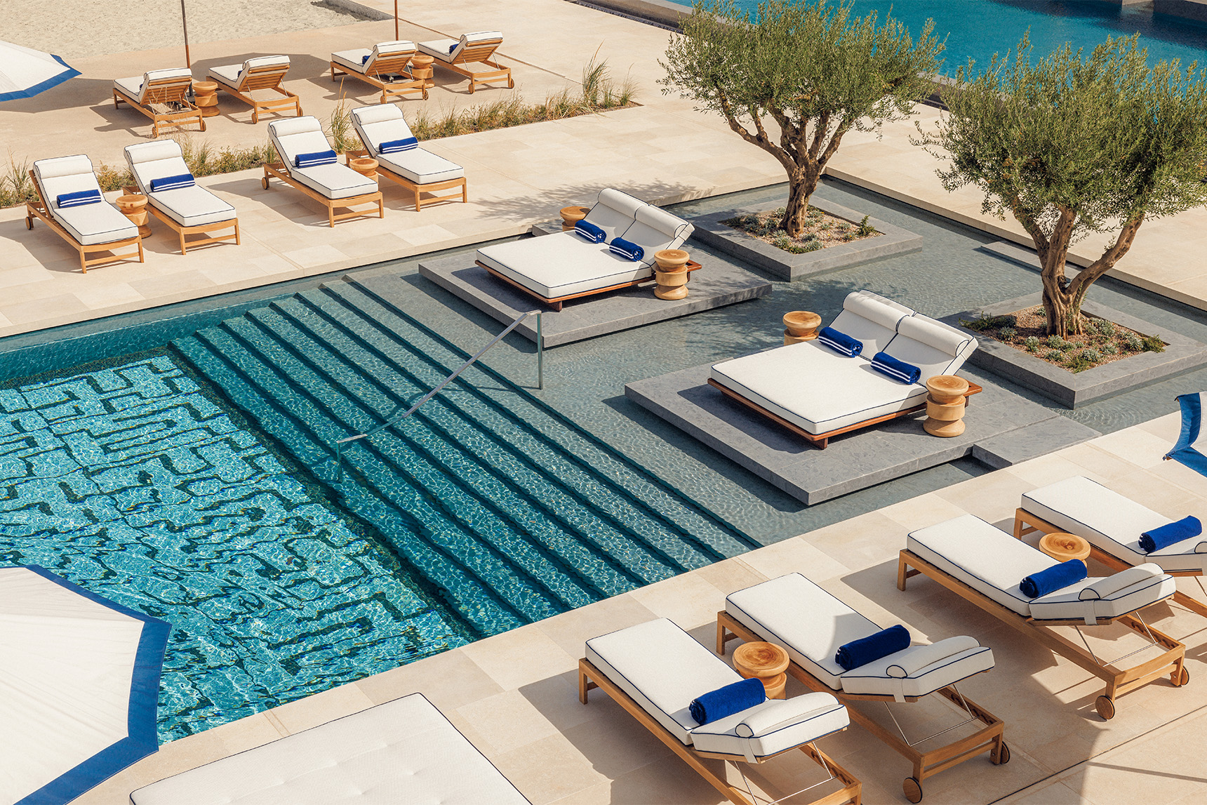 A blue pool with steps down is surrounded by white sun loungers on a sandy coloured patio