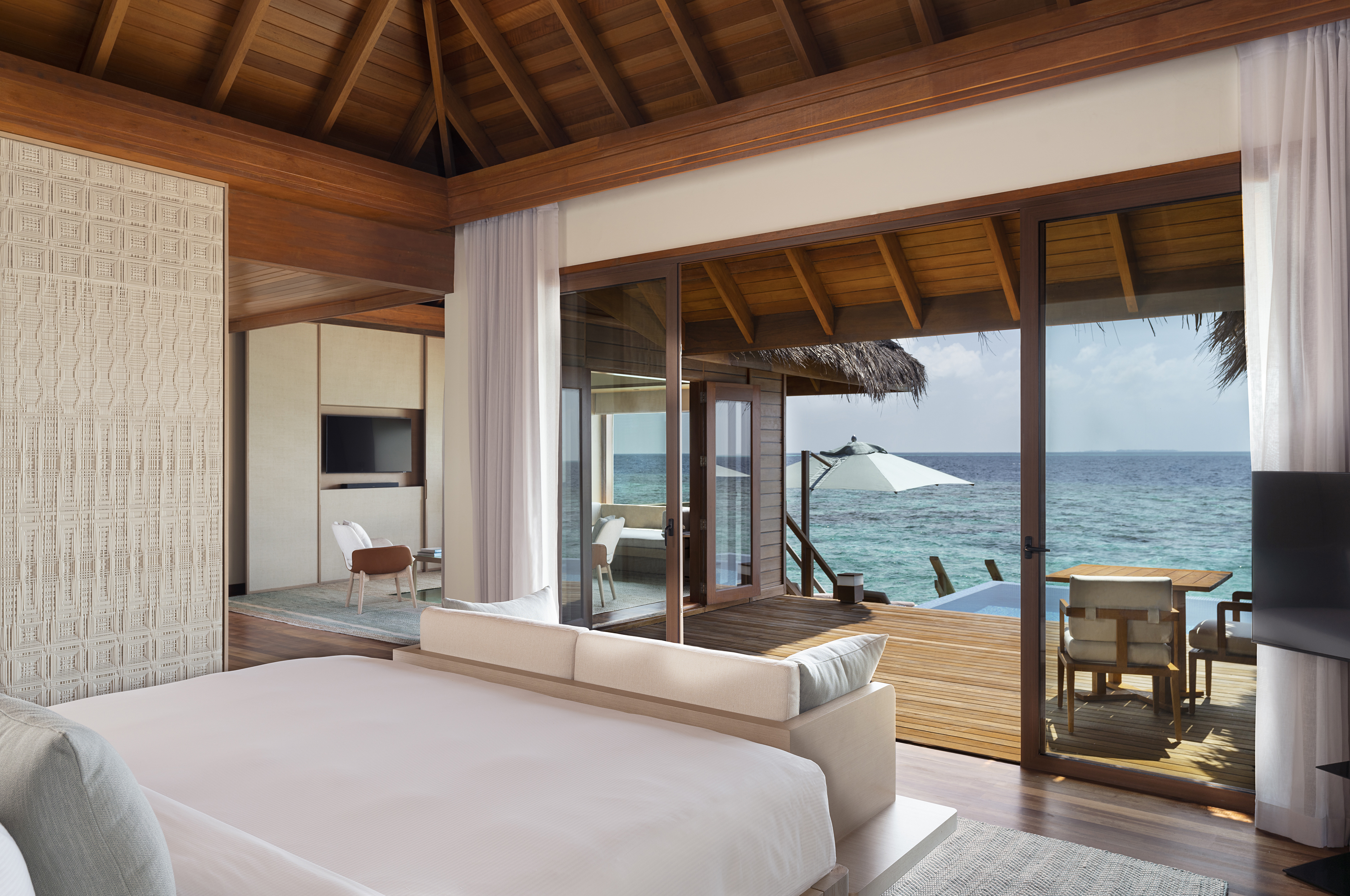 The bedroom of an Ocean Bungalow at Huvafen Fushi featuring the end of a bed looking out towards the ocean with a sofa at the bottom leading onto a deck with pool and bistro table set