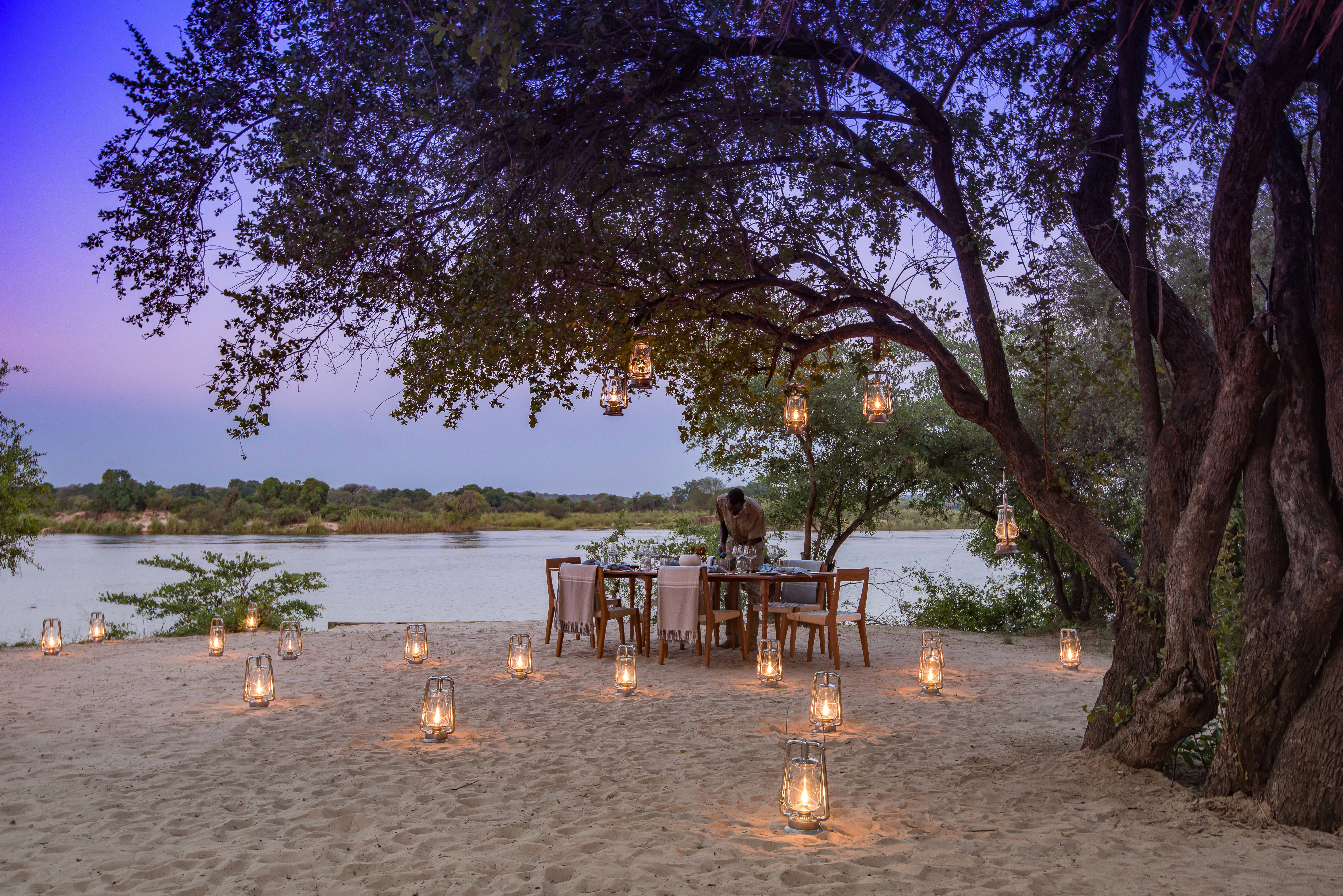 A man setting up an outdoor dining experience beneath a tree on a sandy riverbank with lanterns