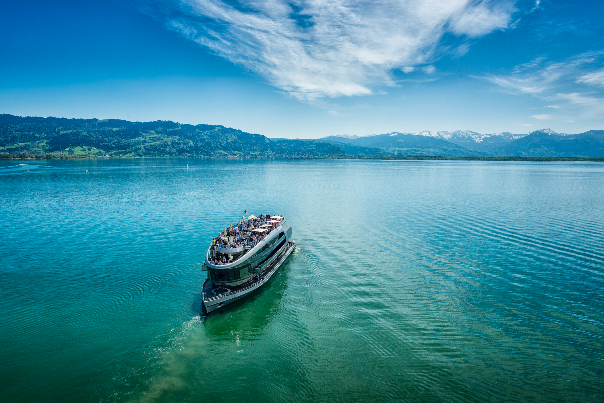 Large passenger boat cruising across a clear blue lake with distant mountains and rolling green hills under a bright sky.
