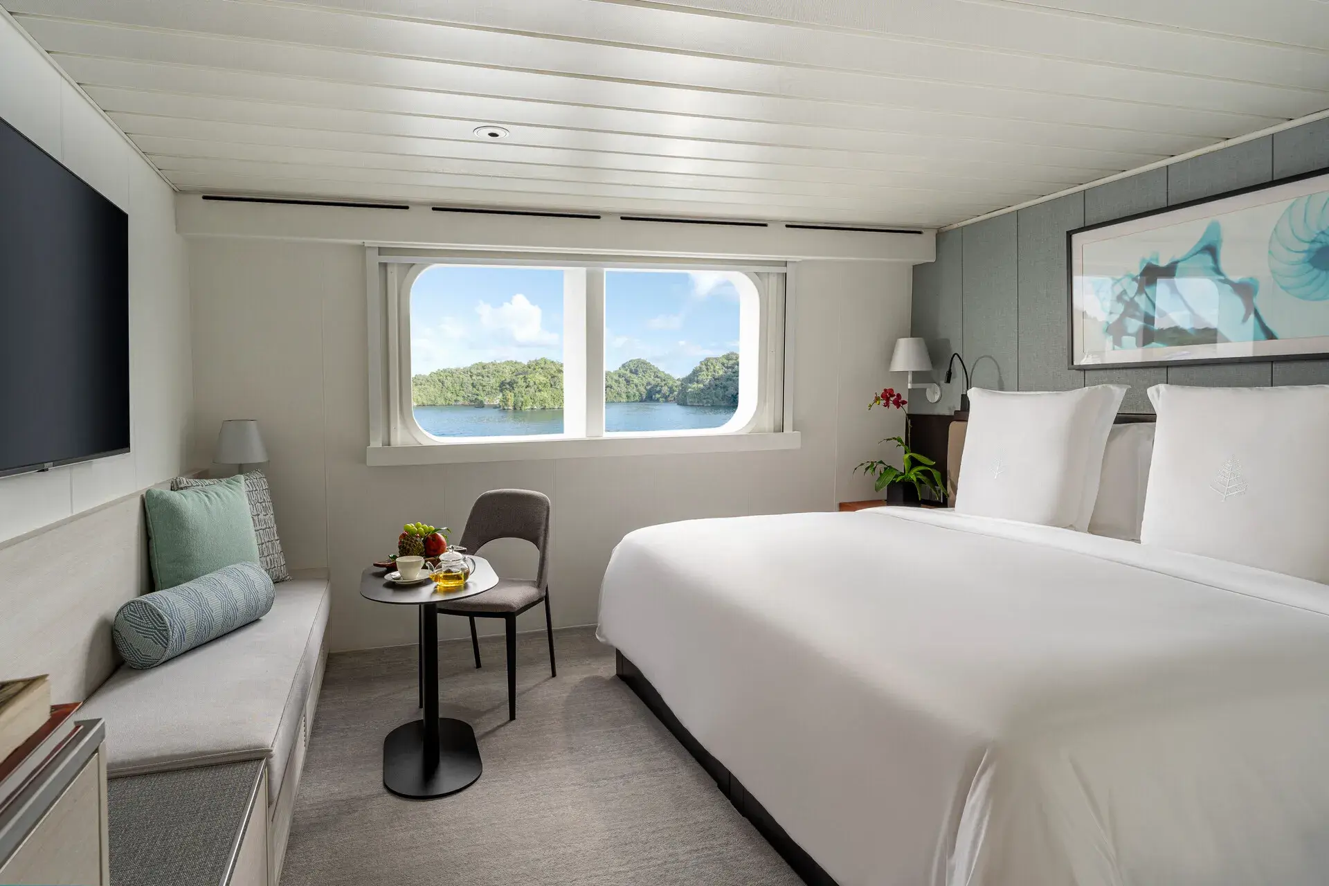 A stylish State Room aboard Four Seasons Explorer Palau featuring decorated in pale colours, with a comfy bed, small lounge area and a window looking out islands beyond