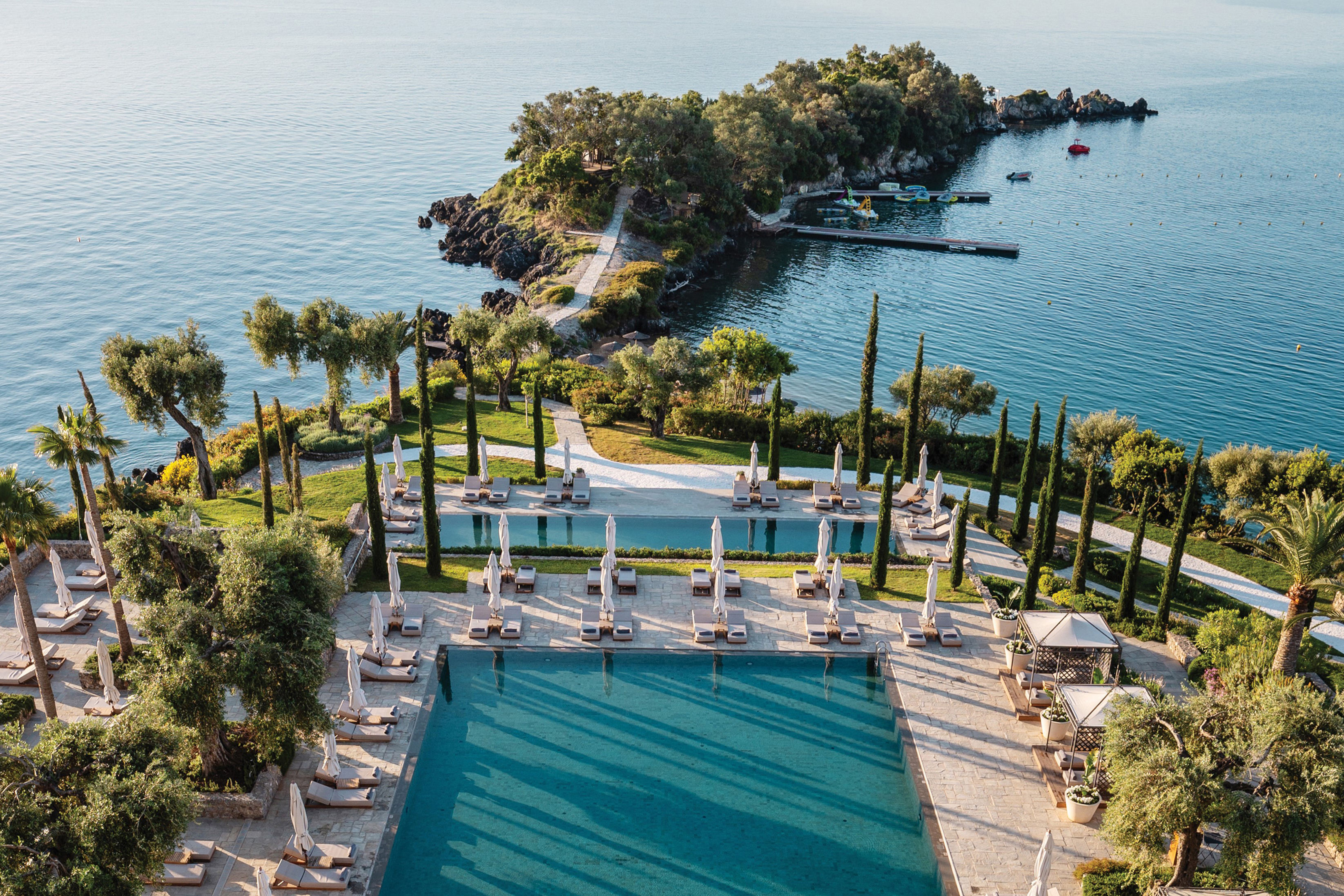 Aerial view of Grecotel Corfu Imperial’s luxury pool area with sun loungers, lush greenery, and Ionian Sea views.