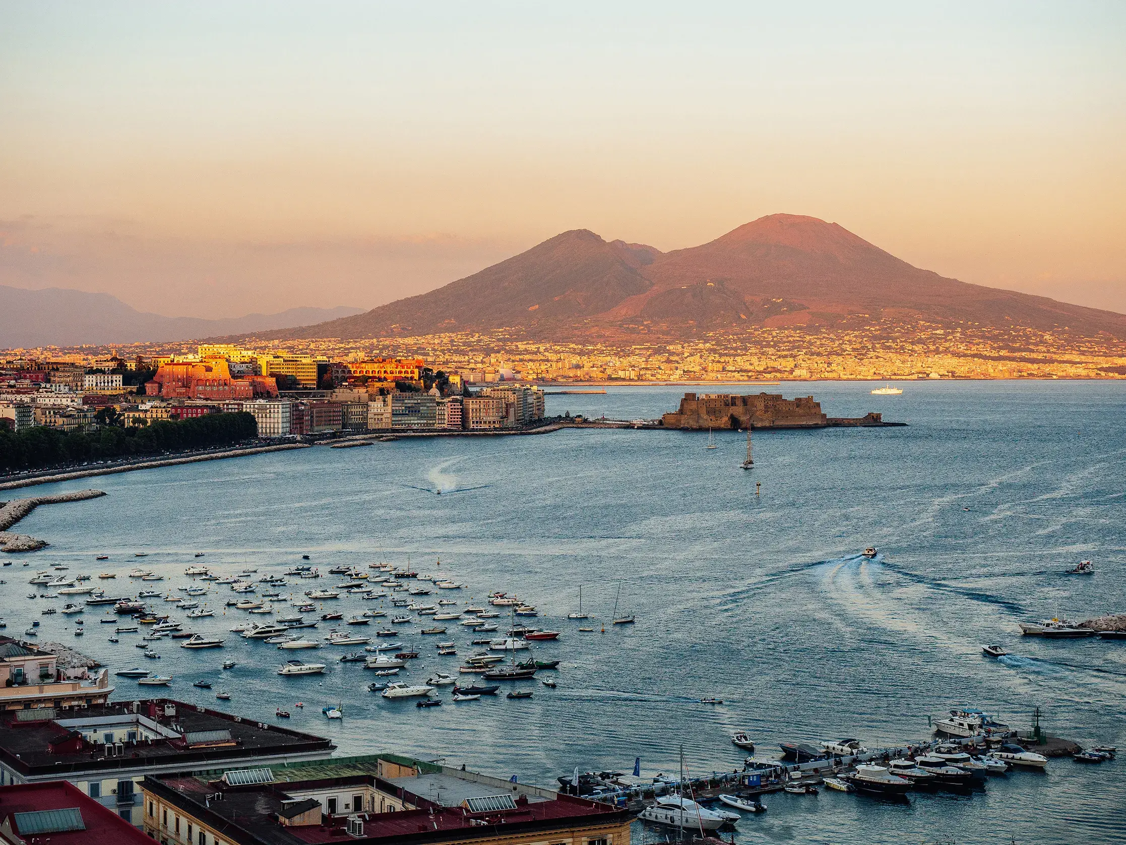 Panoramic view of Naples waterfront with boats in the bay and Mount Vesuvius in the background at sunset.